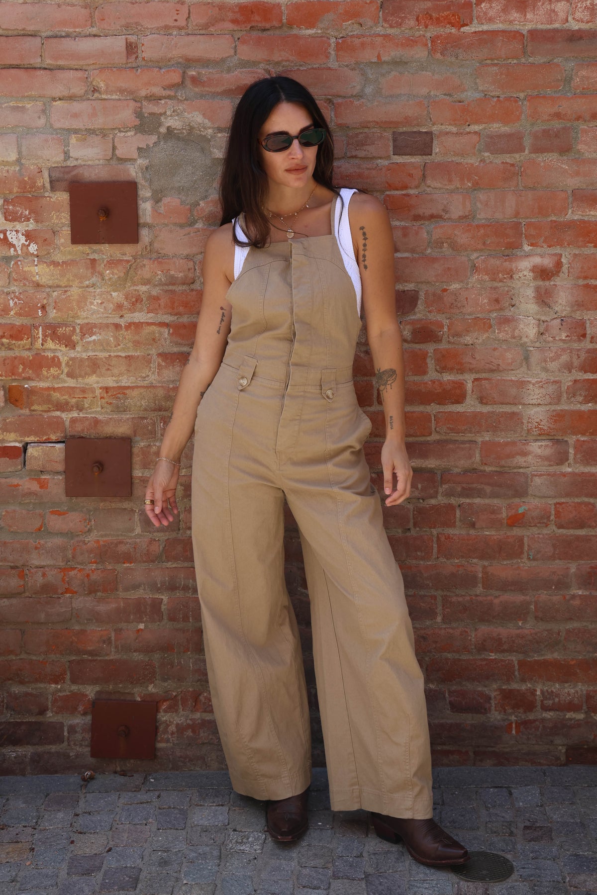 Woman wearing beige overalls standing against a brick wall.