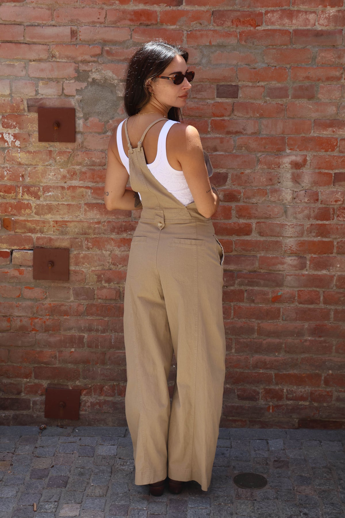 Woman wearing beige overalls and sunglasses against a brick wall.