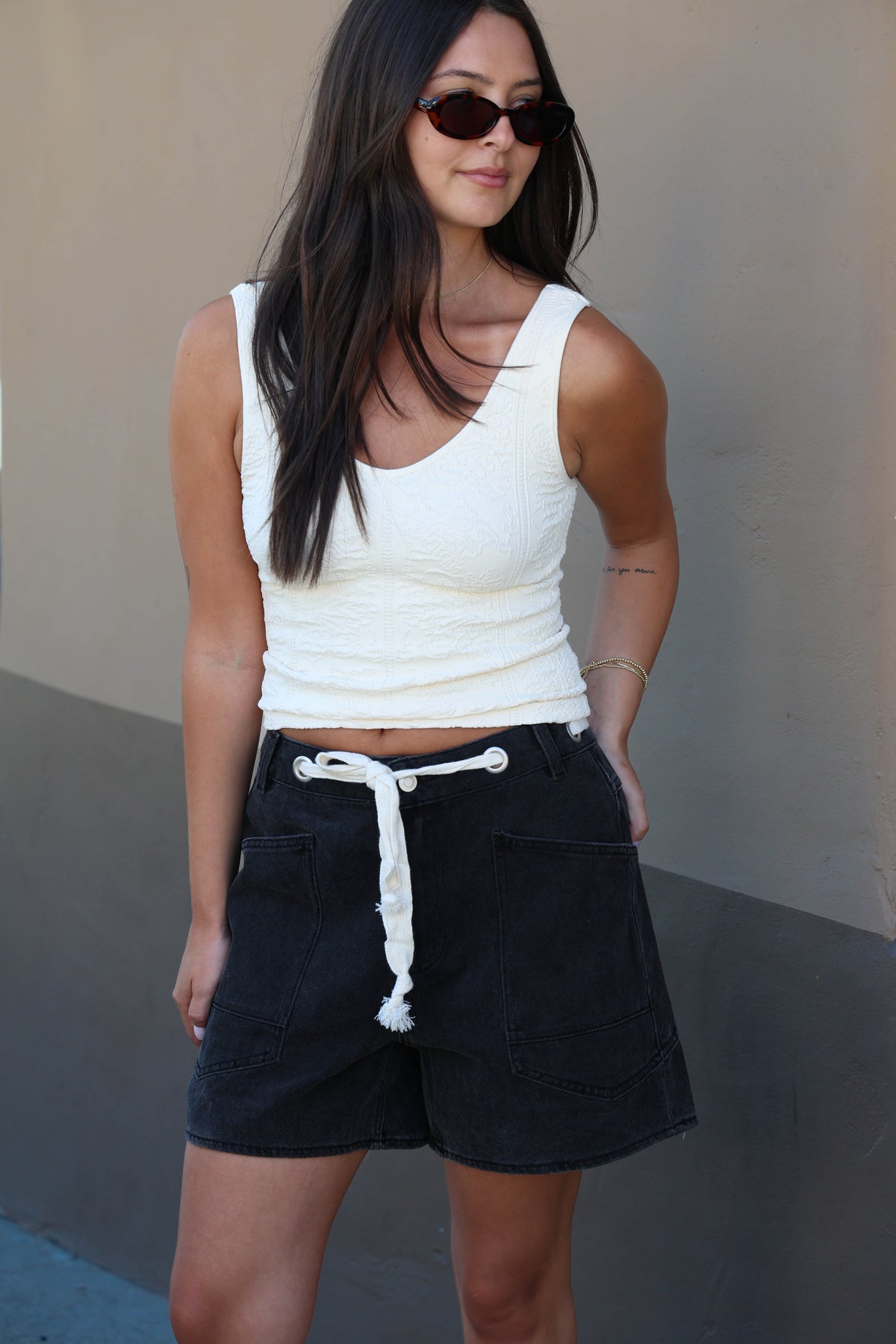 A woman wearing a white ribbed tank top and dark denim shorts with a white drawstring, standing against a neutral gray wall.
