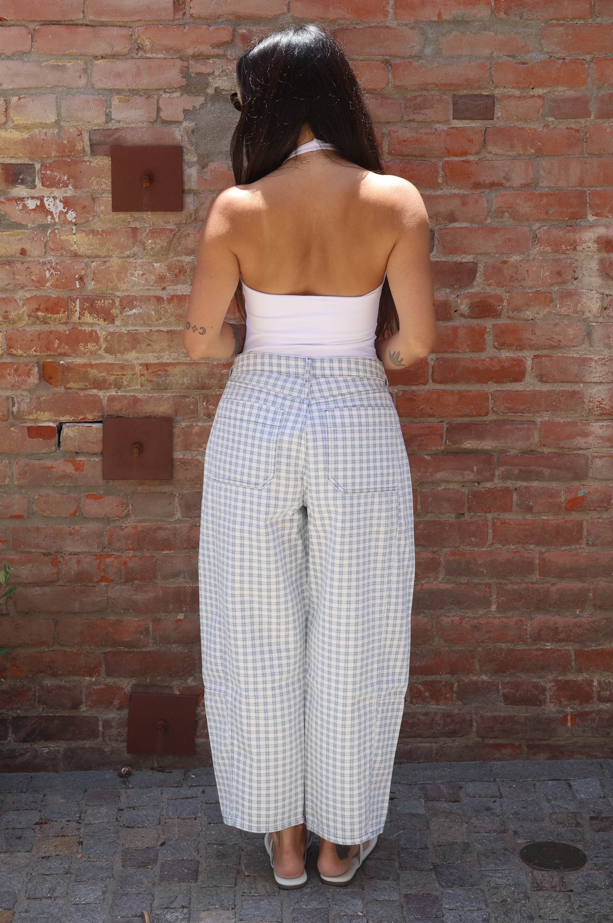 Woman wearing a white top and checkered pants standing against a brick wall.
