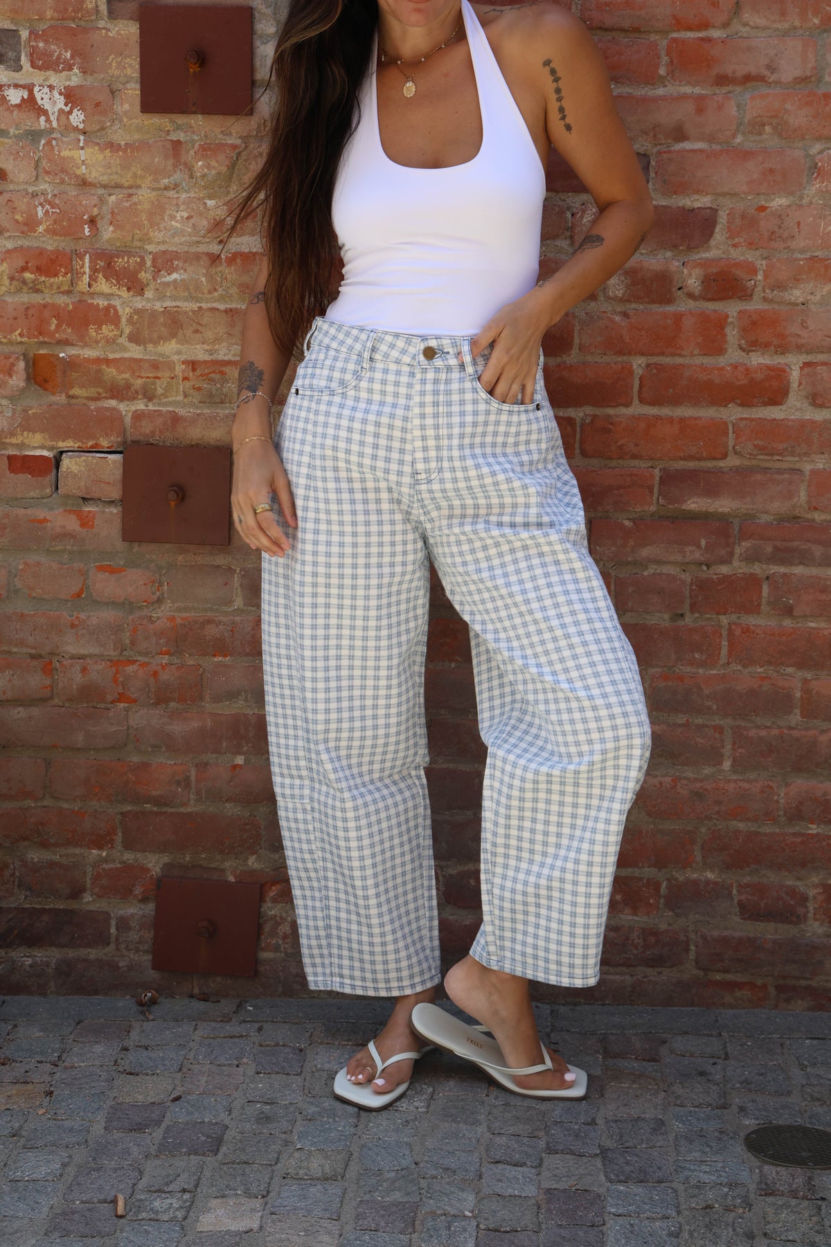 Person wearing a white tank top and checkered pants standing against a brick wall.