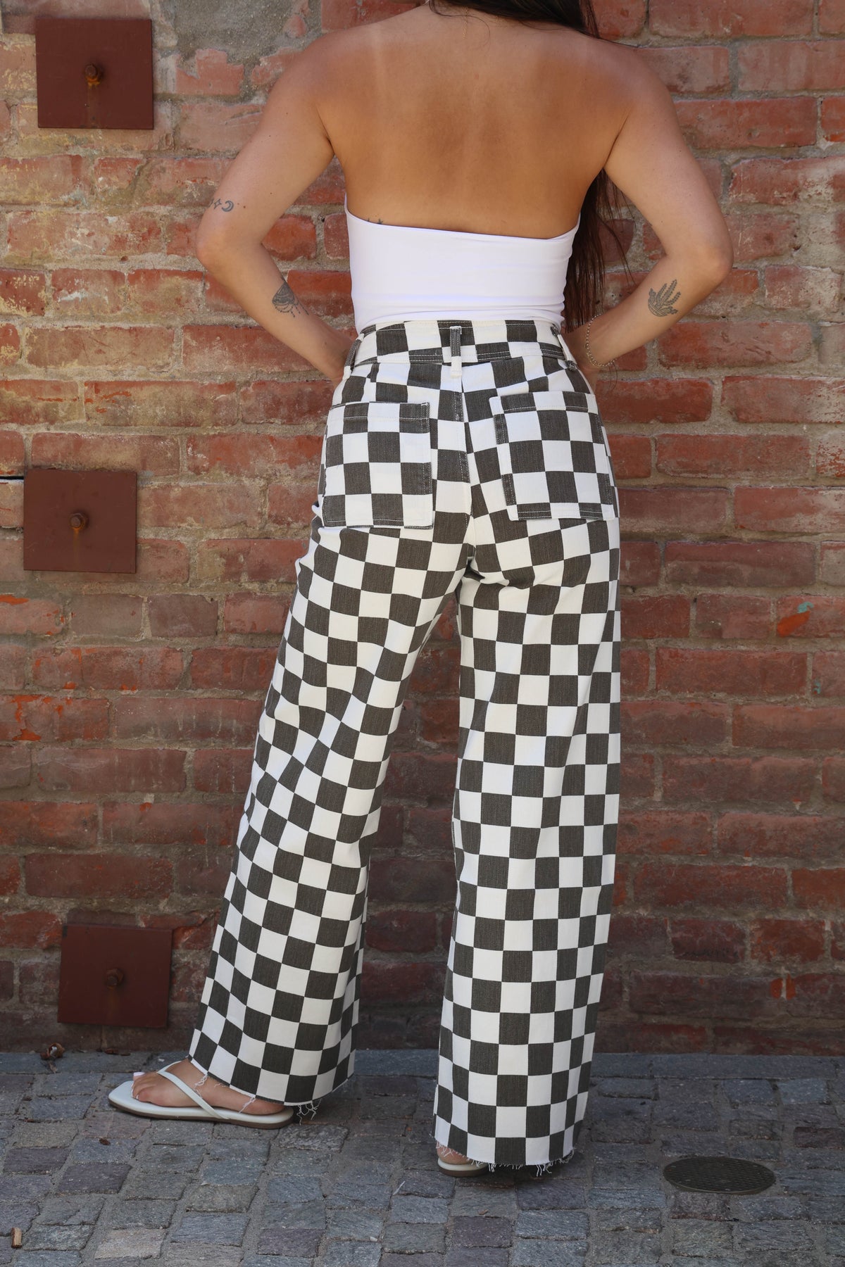 Person wearing a white top and black and white checkered pants standing against a brick wall.