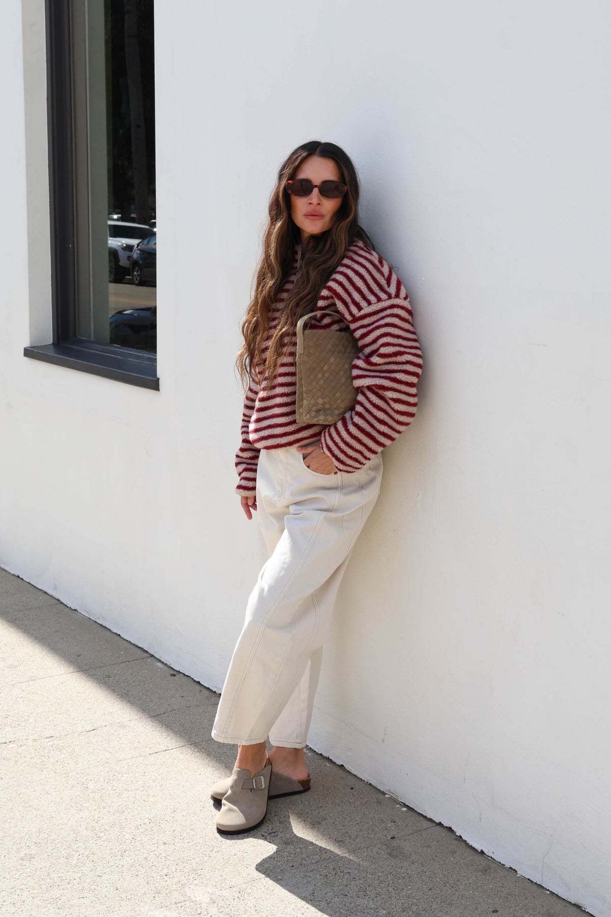Woman wearing a striped cream and red fleece, sunglasses and white jeans holding a bag.