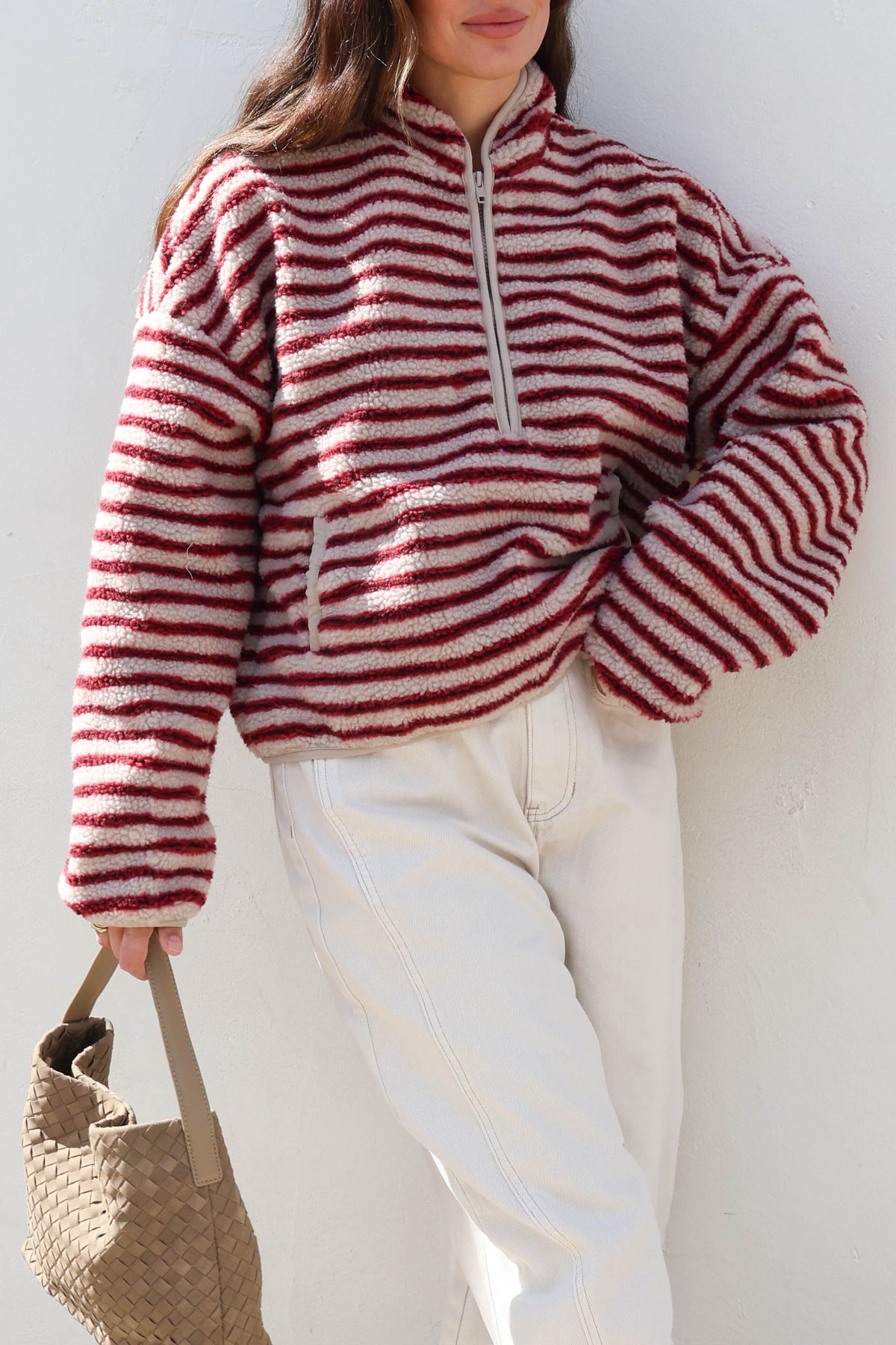 Woman wearing a striped cream and red fleece, sunglasses and white jeans holding a bag.