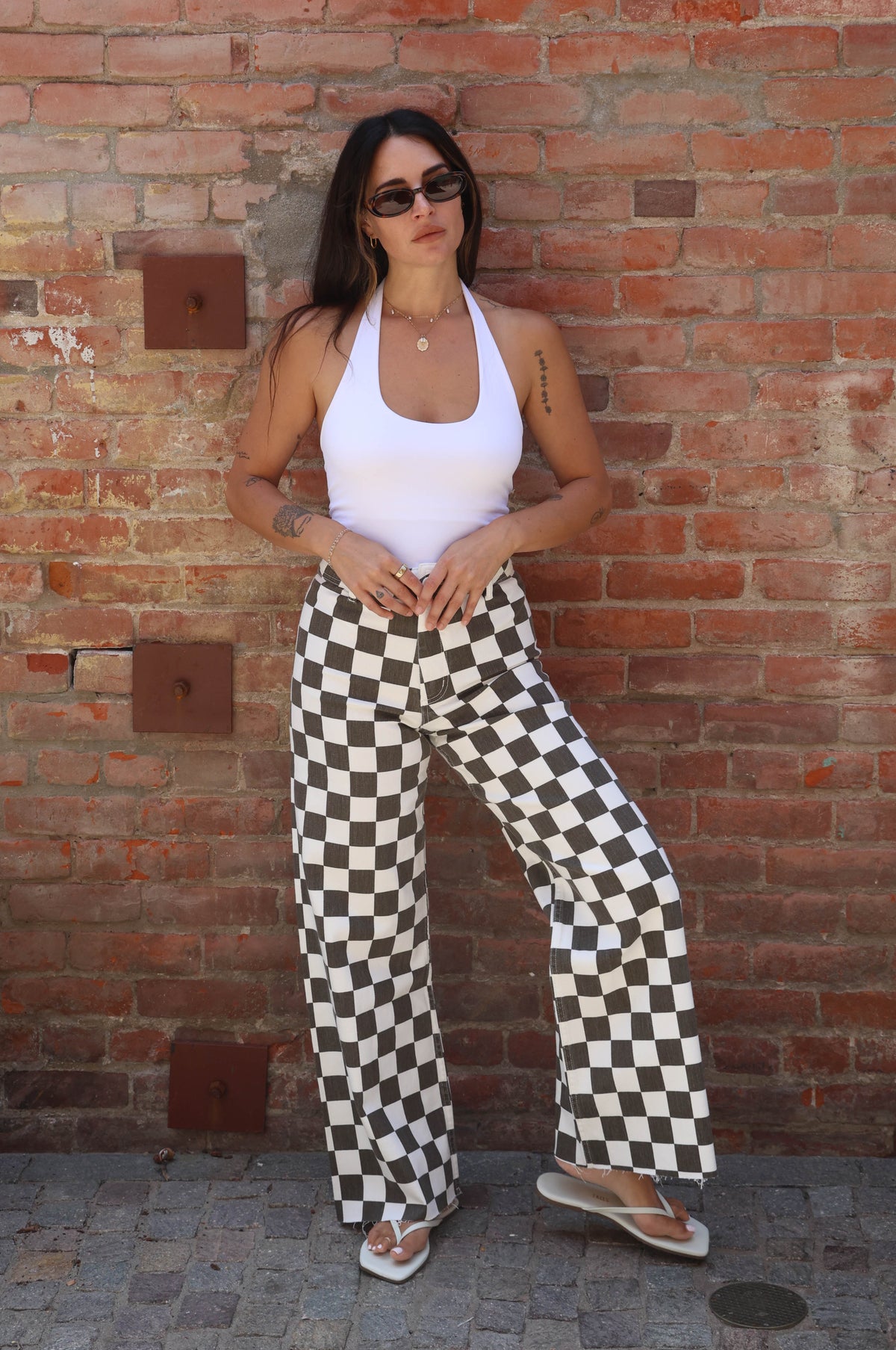 Woman wearing a white top and checkered pants standing against a brick wall.