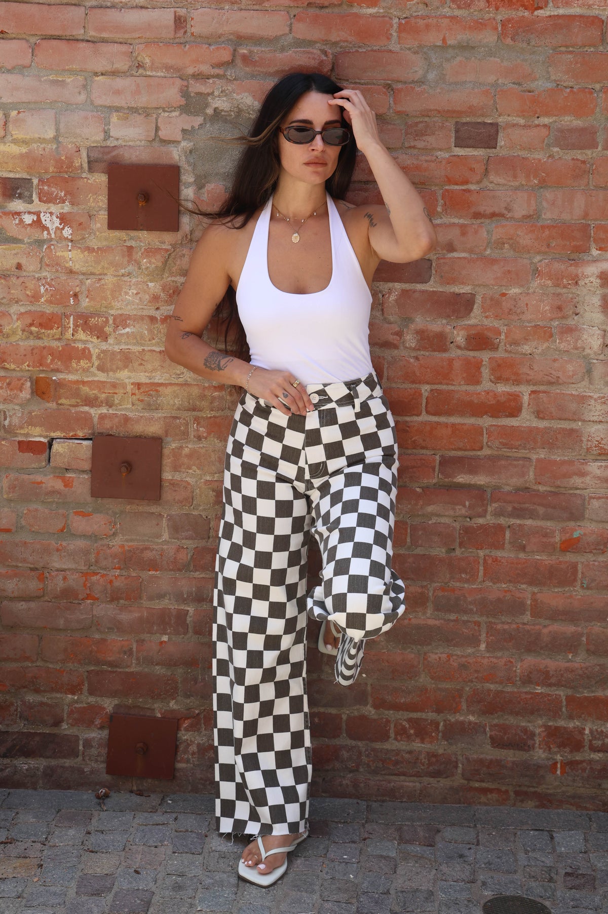Woman wearing a white top and checkered pants against a brick wall.