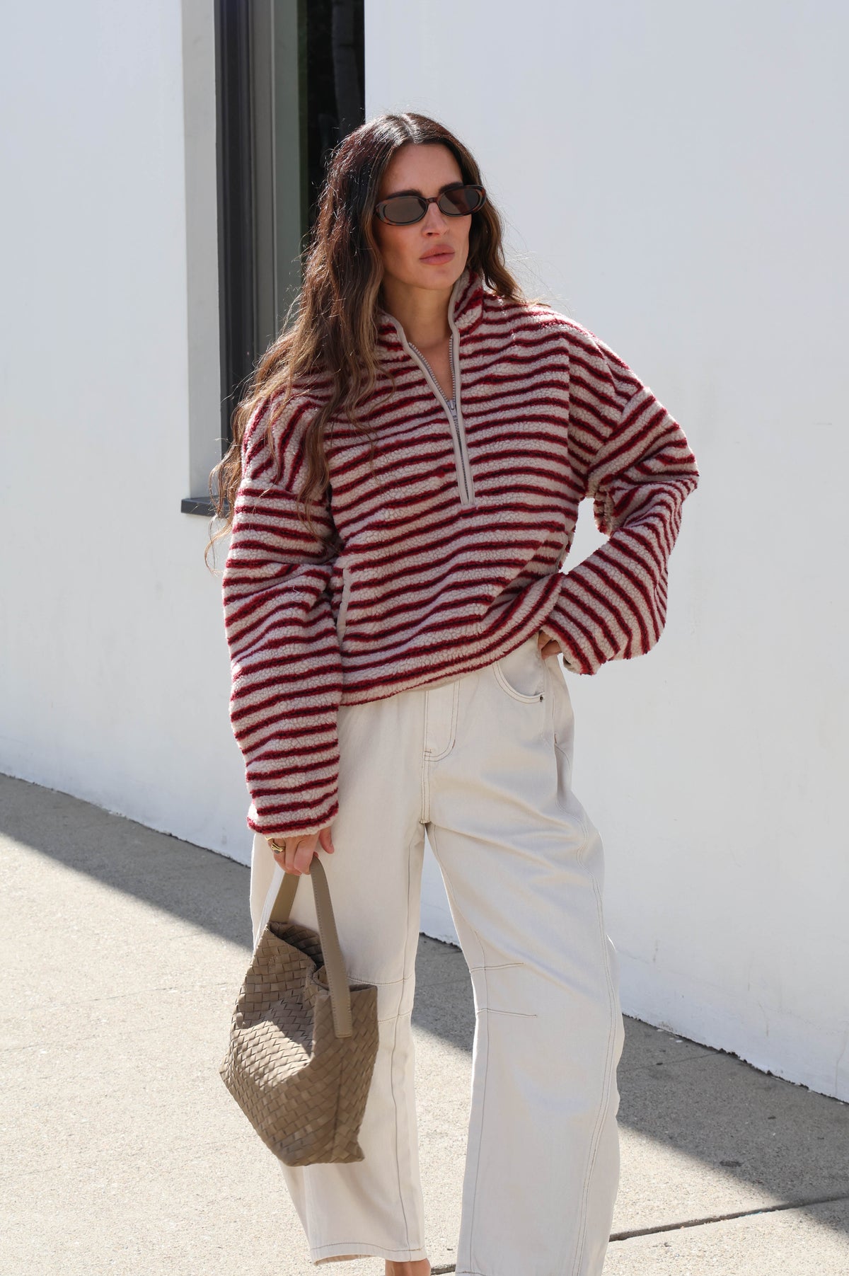 Woman wearing a striped cream and red fleece, sunglasses and white jeans holding a bag.