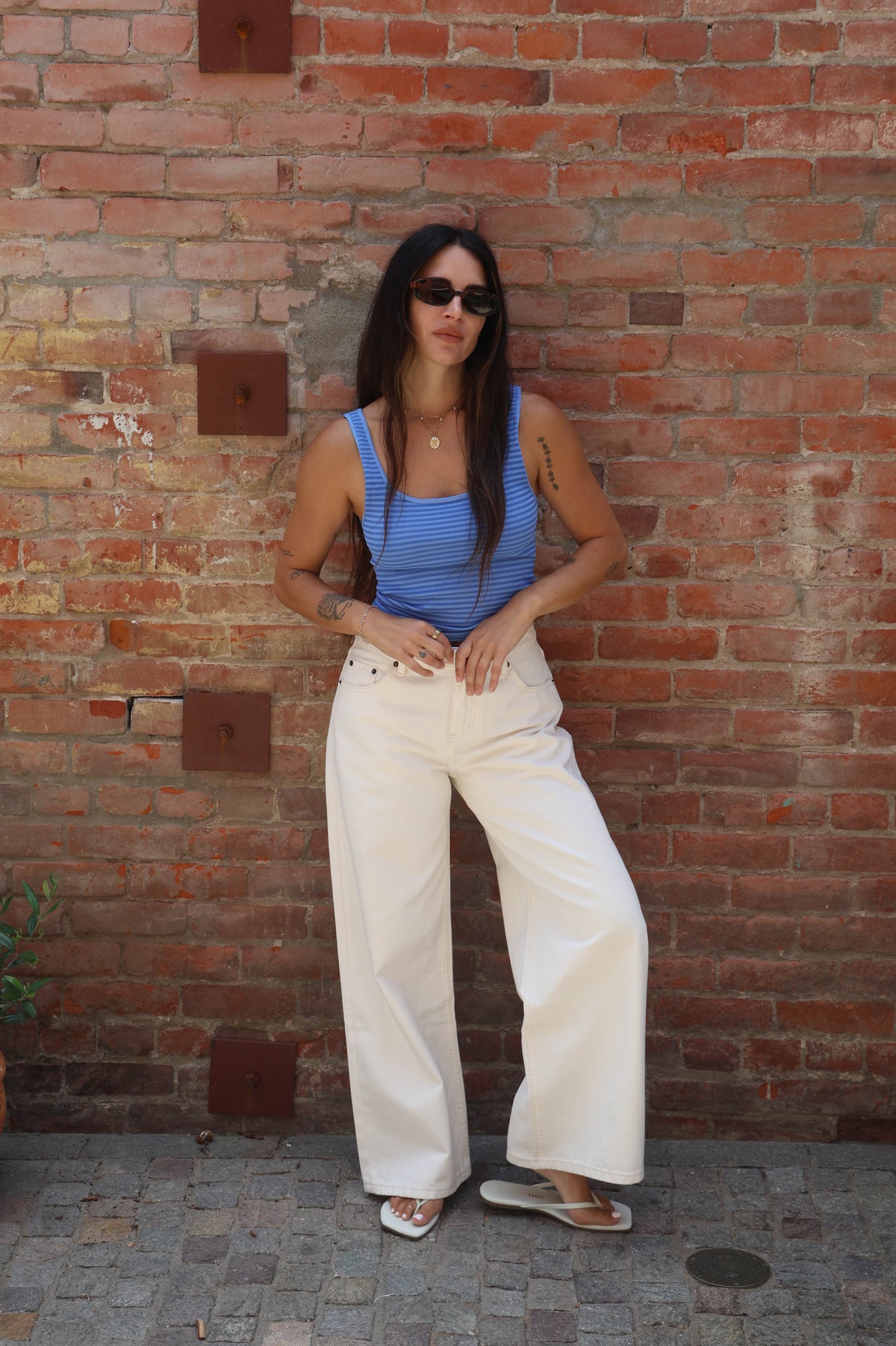 Woman in blue top and white pants standing against a brick wall.