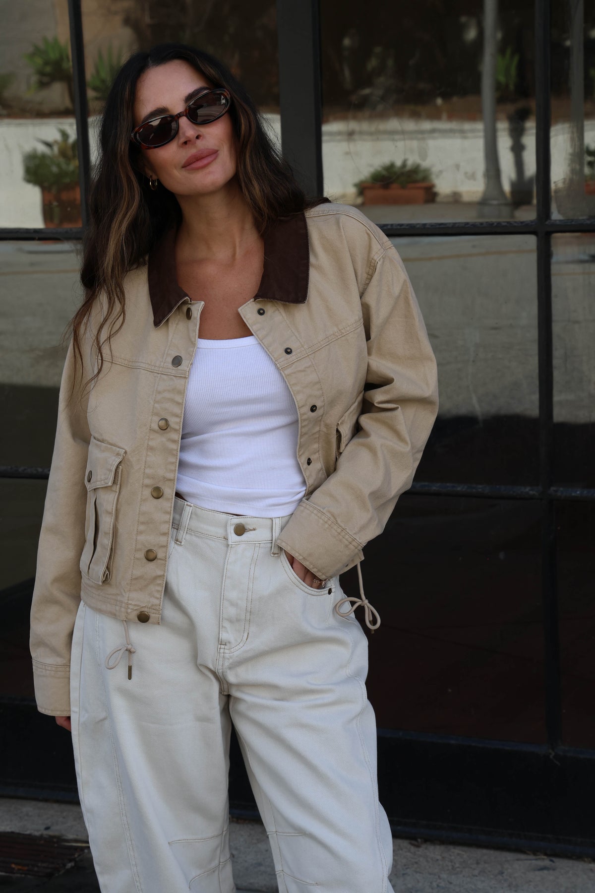 Model is wearing a brown canvas jacket with a white tank, white jeans and sunglasses.