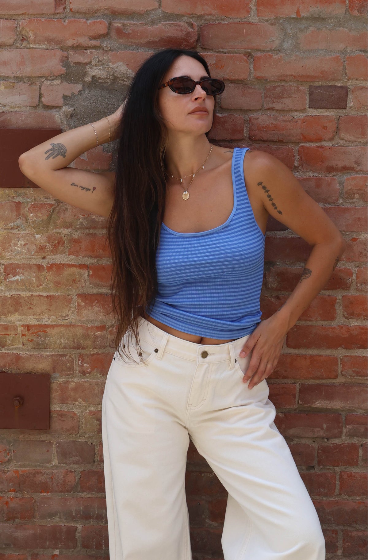 Woman in blue top and white pants standing against a brick wall.