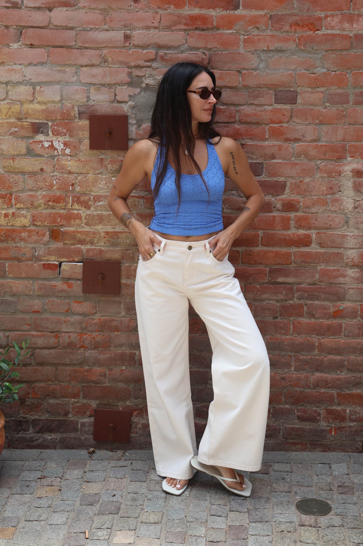 Woman in blue top and white pants standing against a brick wall.