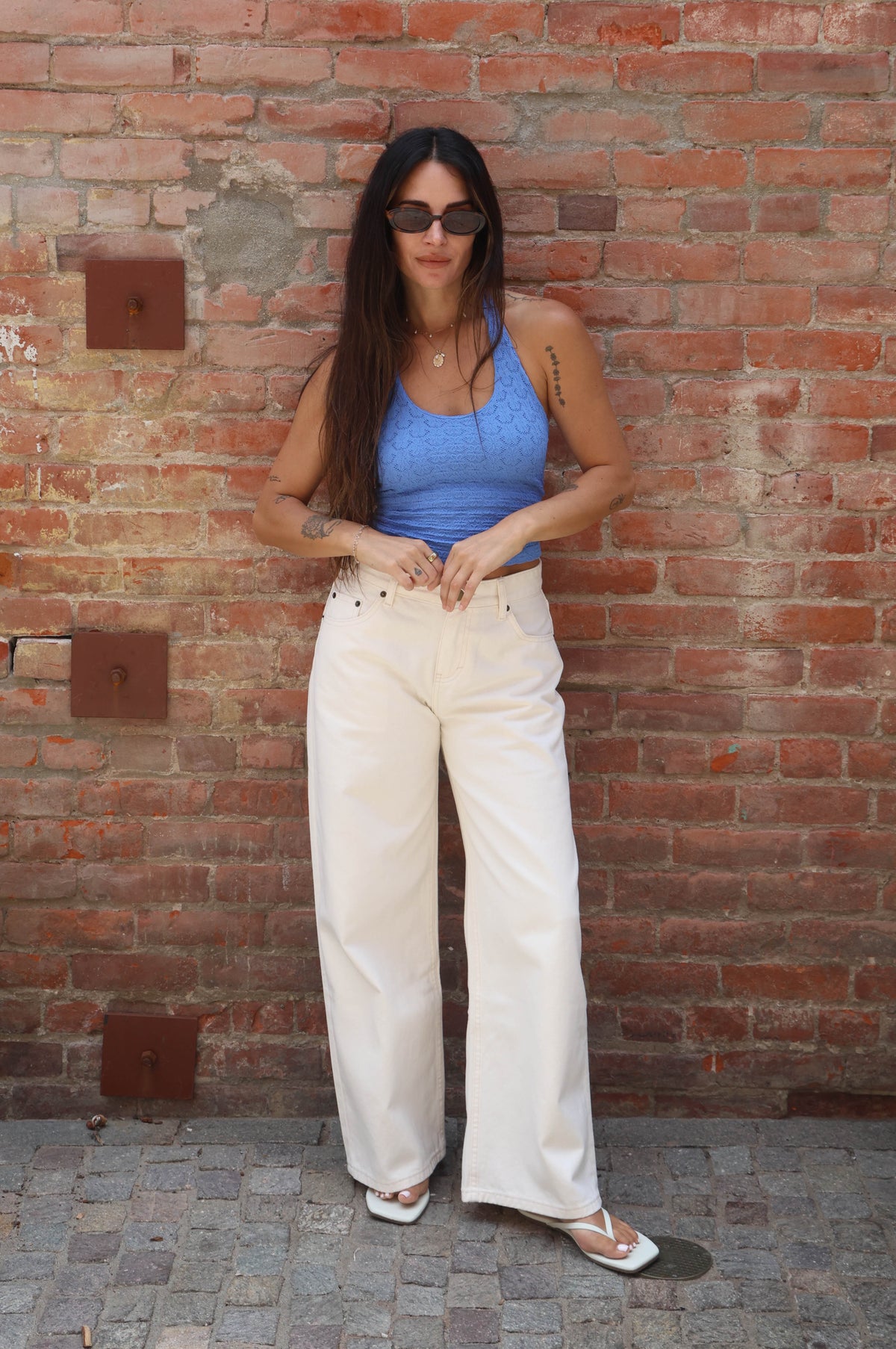Woman in a blue top and white pants standing against a brick wall.