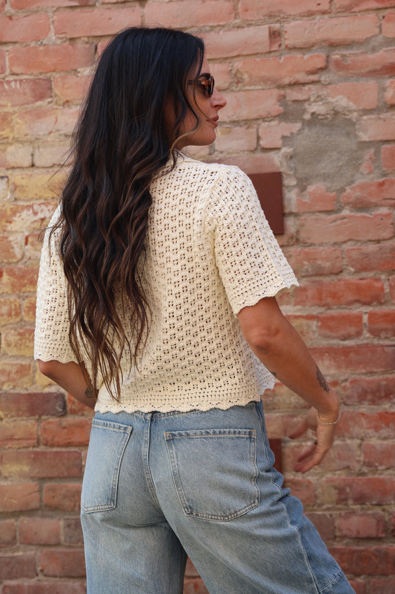 Woman wearing a cream-colored crochet cardigan with jeans against a brick wall.