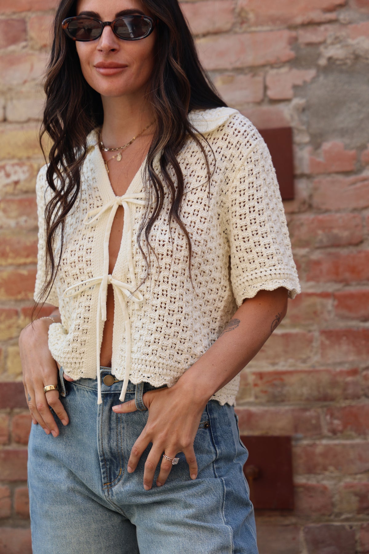 Woman wearing a cream-colored crochet cardigan with jeans against a brick wall.
