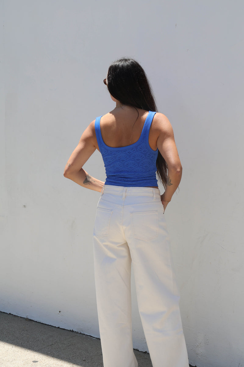 Person wearing a blue tank top and white pants against a white wall