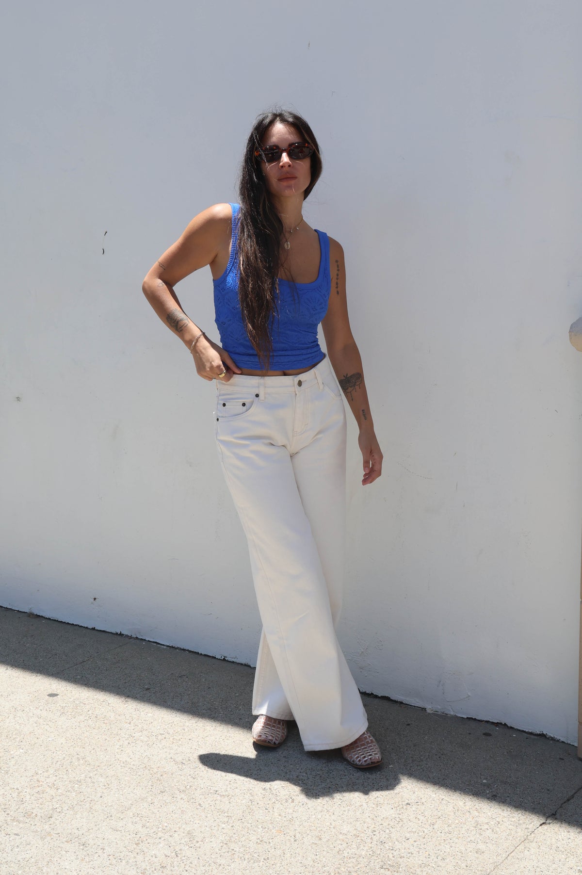 Woman wearing a blue top and white pants standing against a white wall.