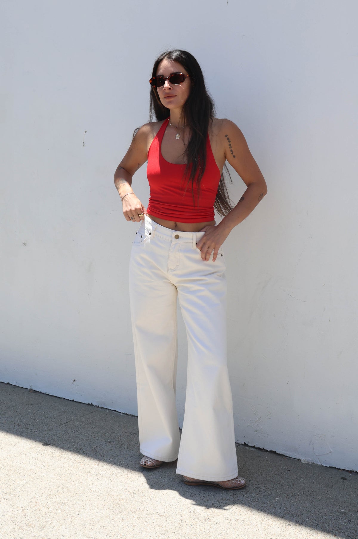 Woman wearing a red top and white pants standing against a white wall.