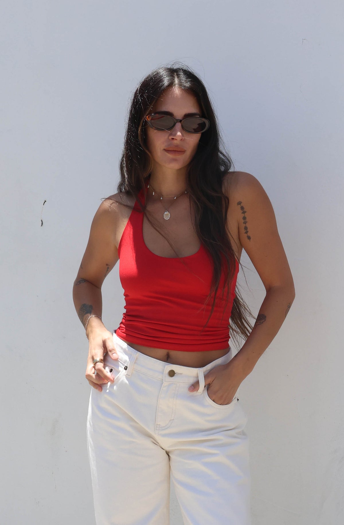 Woman wearing a red top and white pants standing against a white wall.