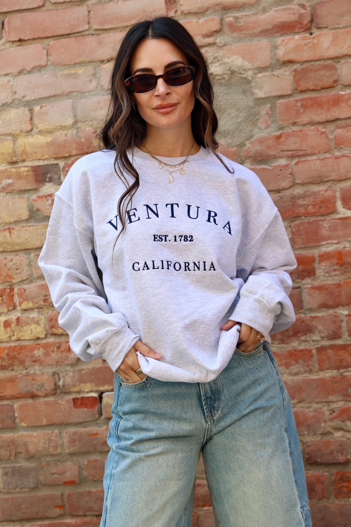 Woman wearing a white sweatshirt with 'Ventura California' text against a brick wall.