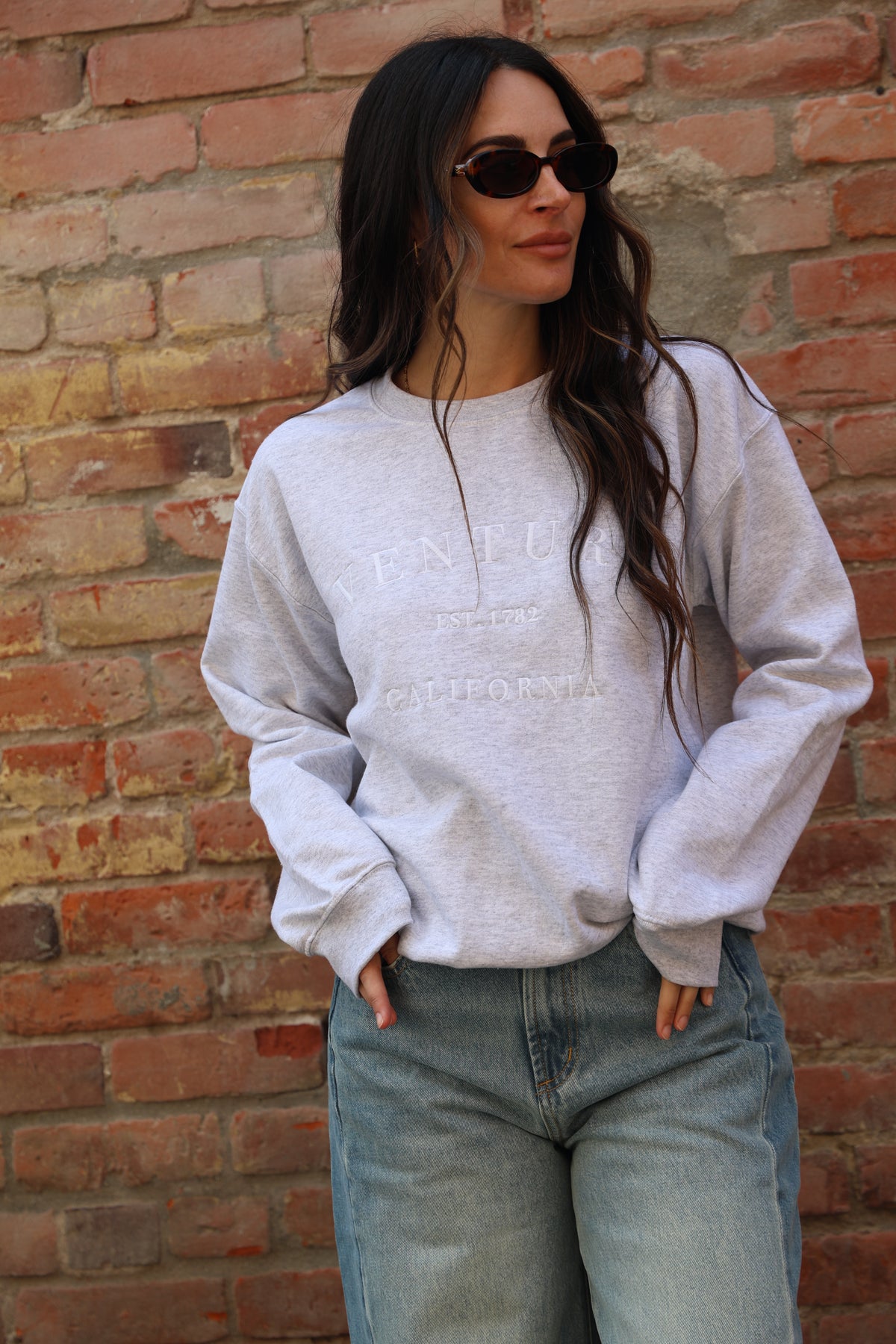 Person wearing a light gray sweatshirt and sunglasses against a brick wall.