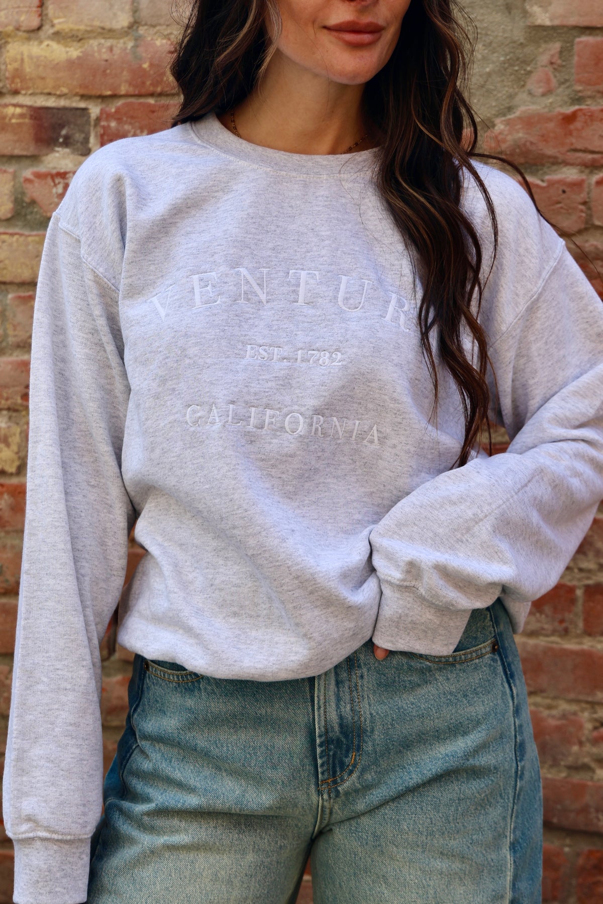 Person wearing a gray sweatshirt with text and blue jeans against a brick wall.