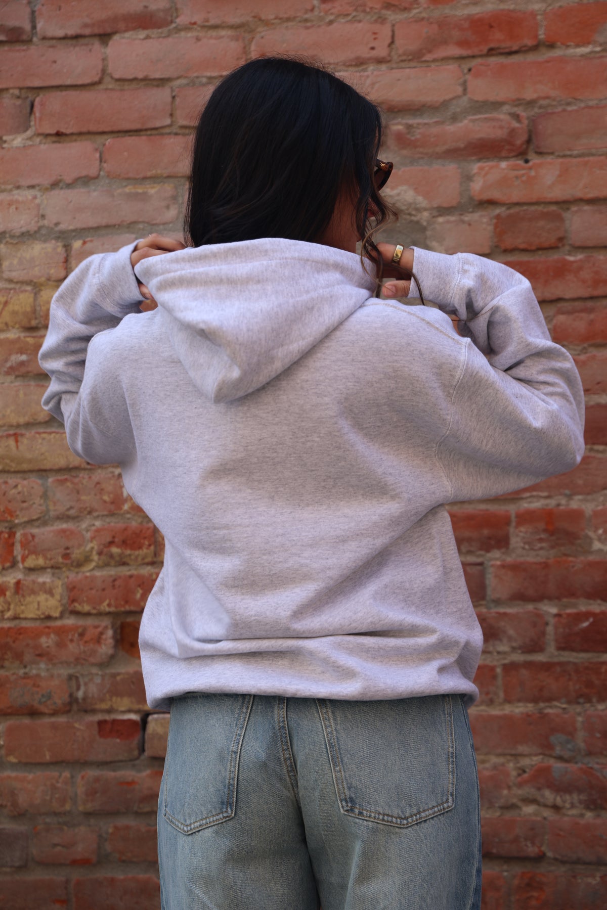 Person wearing a light gray hoodie and blue jeans against a brick wall.