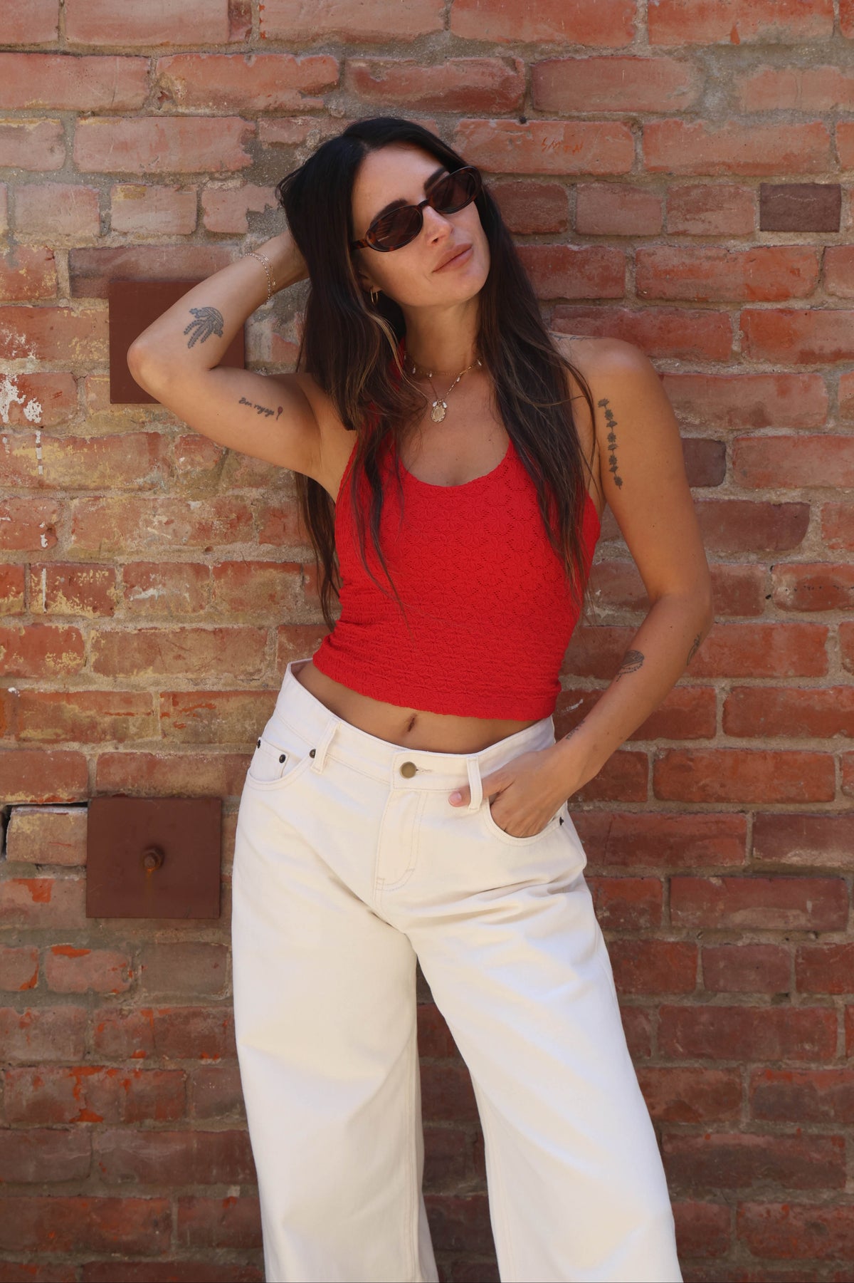 Woman in a red top and white pants standing against a brick wall.