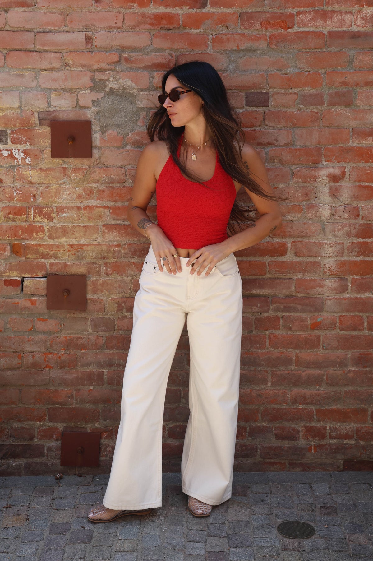 Woman in red top and white pants standing against a brick wall.