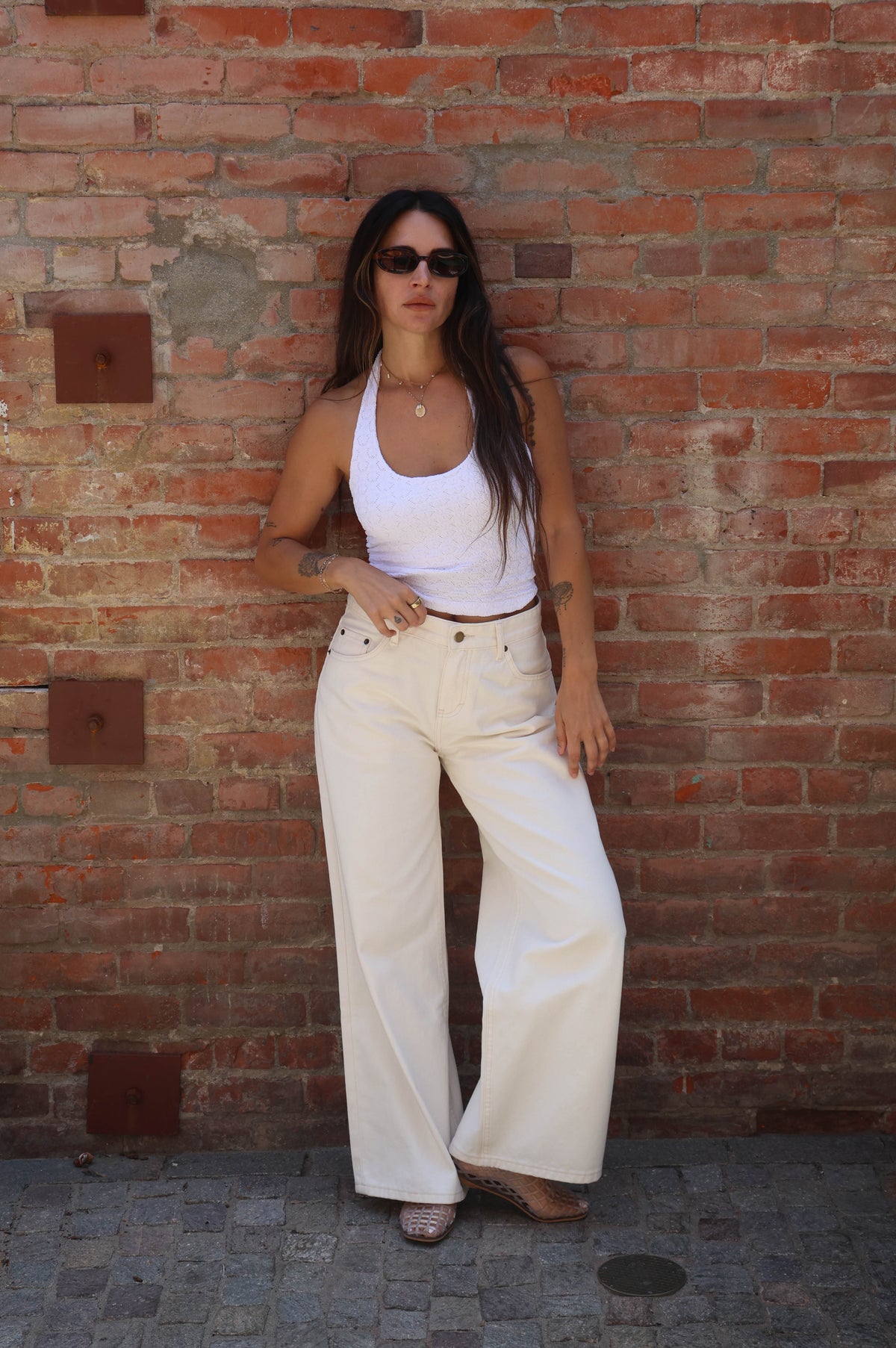 Woman in white tank top and cream pants standing against a brick wall.