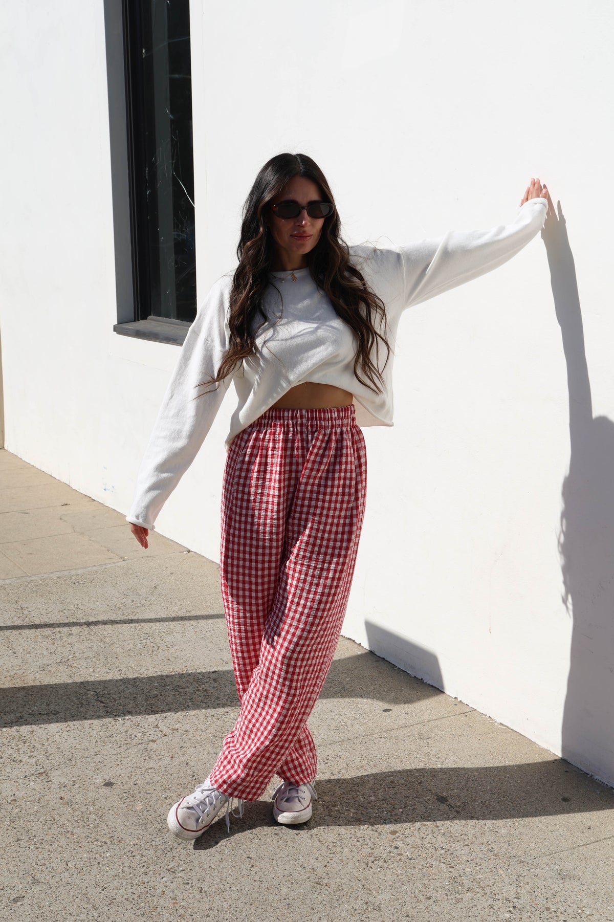 Woman wearing a white sweater and red checkered pants standing against a white wall.