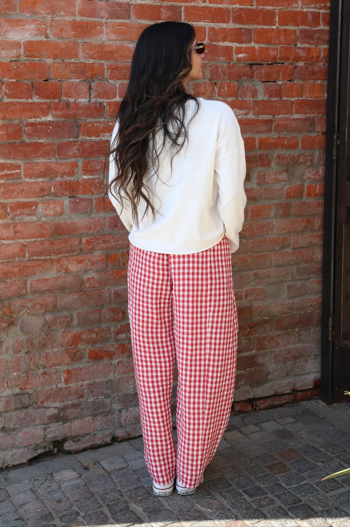 Woman wearing a white sweater and red checkered pants standing against a brick wall.