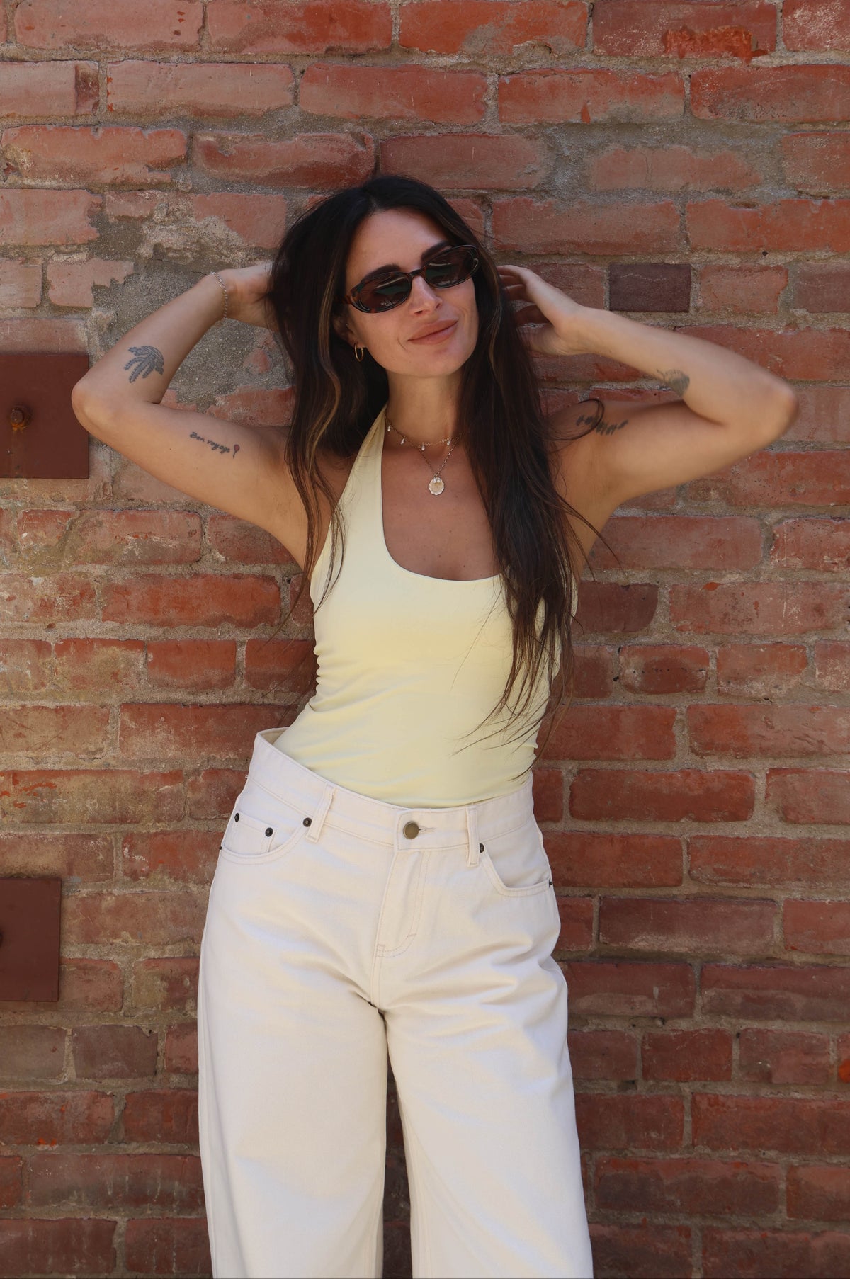 Woman in a yellow top and white pants standing against a brick wall.