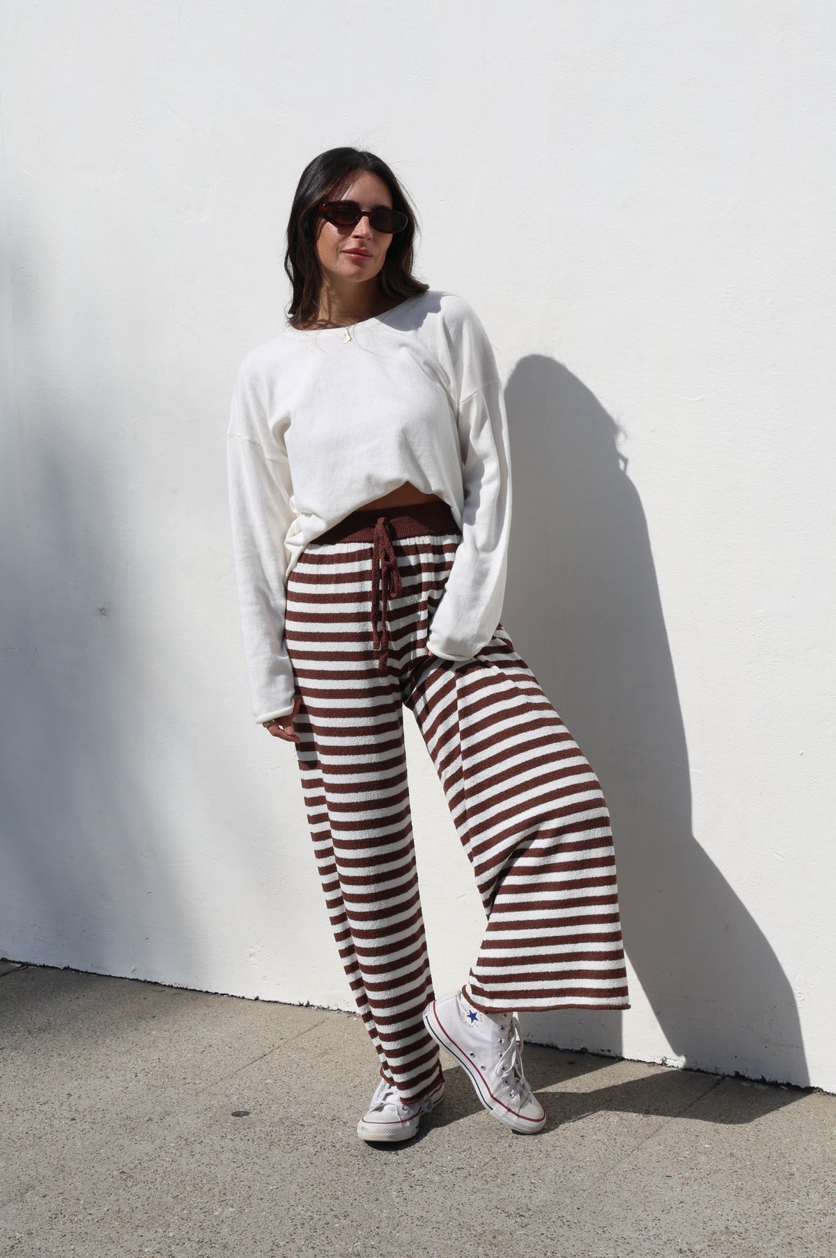 Woman wearing a white sweater and striped pants against a white wall.