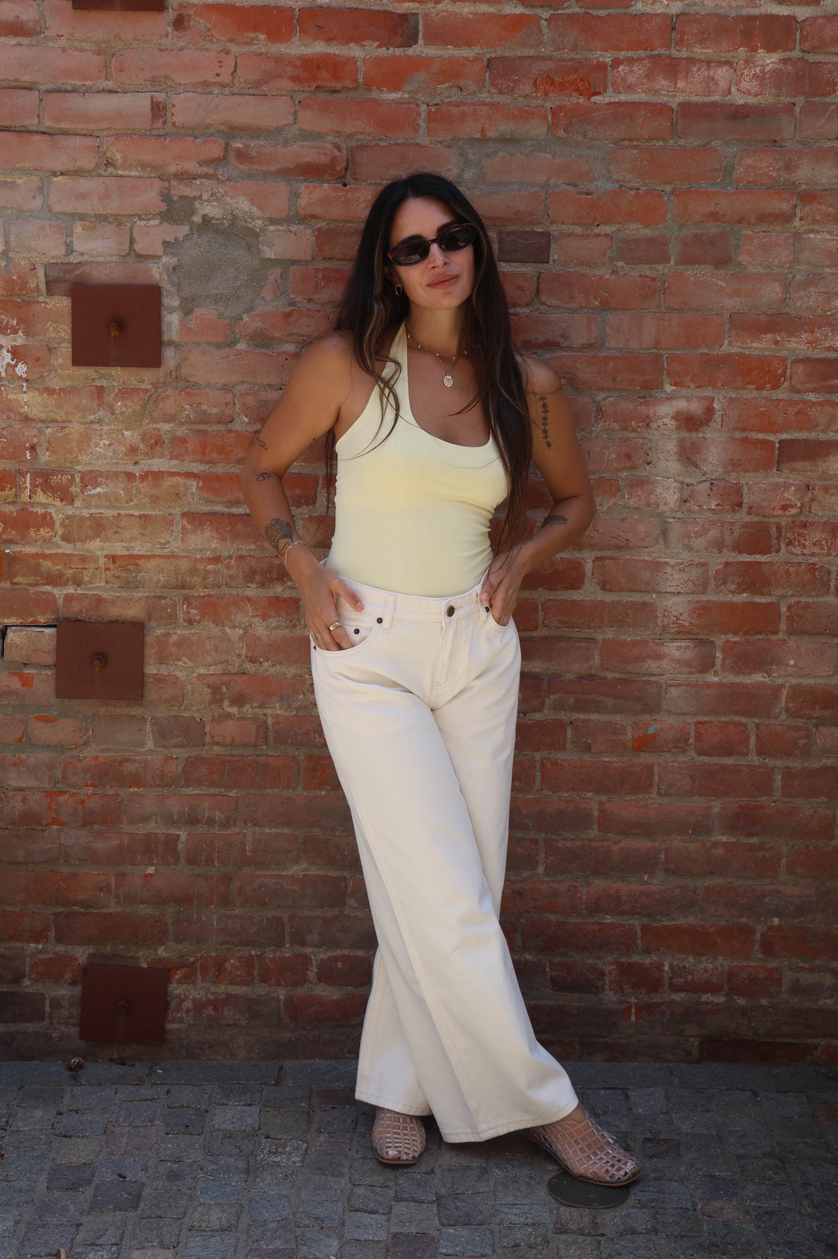Woman in a yellow top and white pants standing against a brick wall.