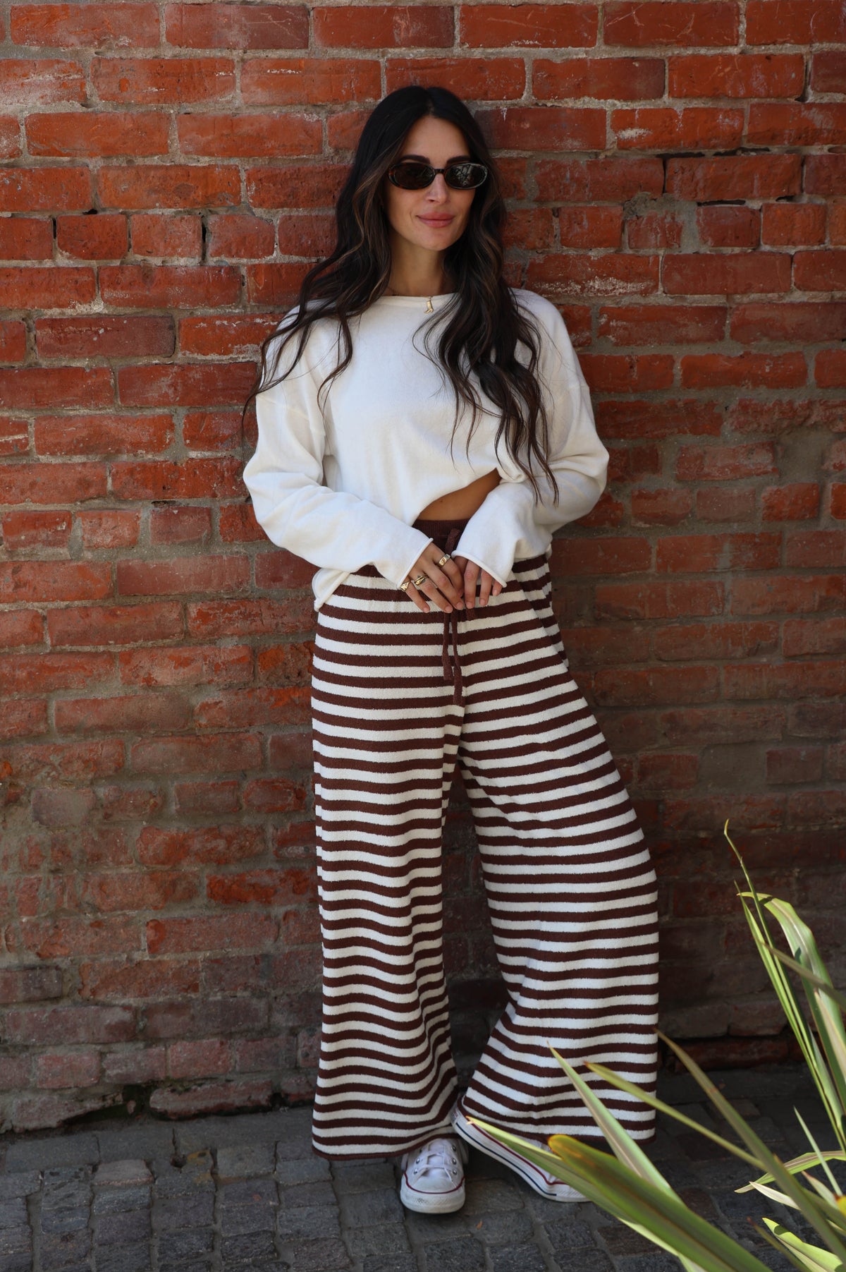 Woman wearing a white top and brown striped pants standing against a brick wall.