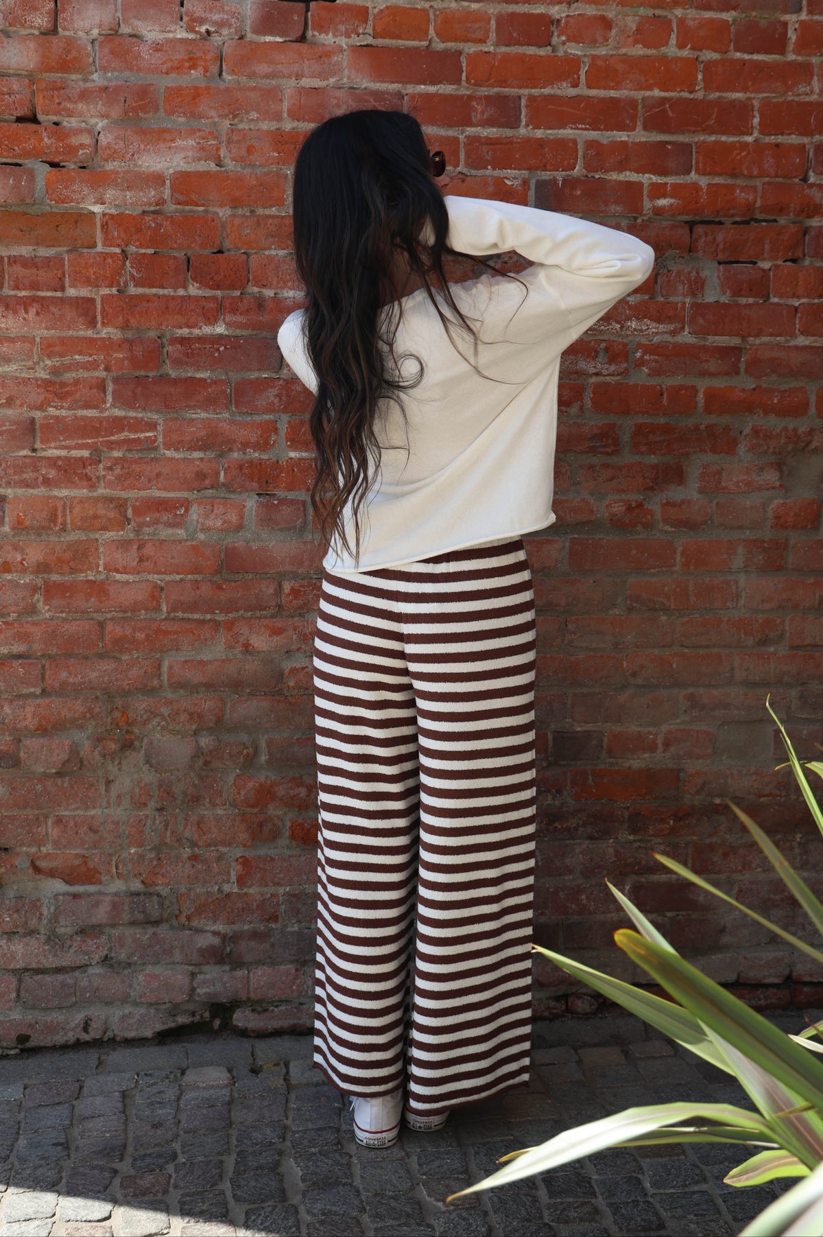 Person wearing a white top and striped pants standing against a brick wall.
