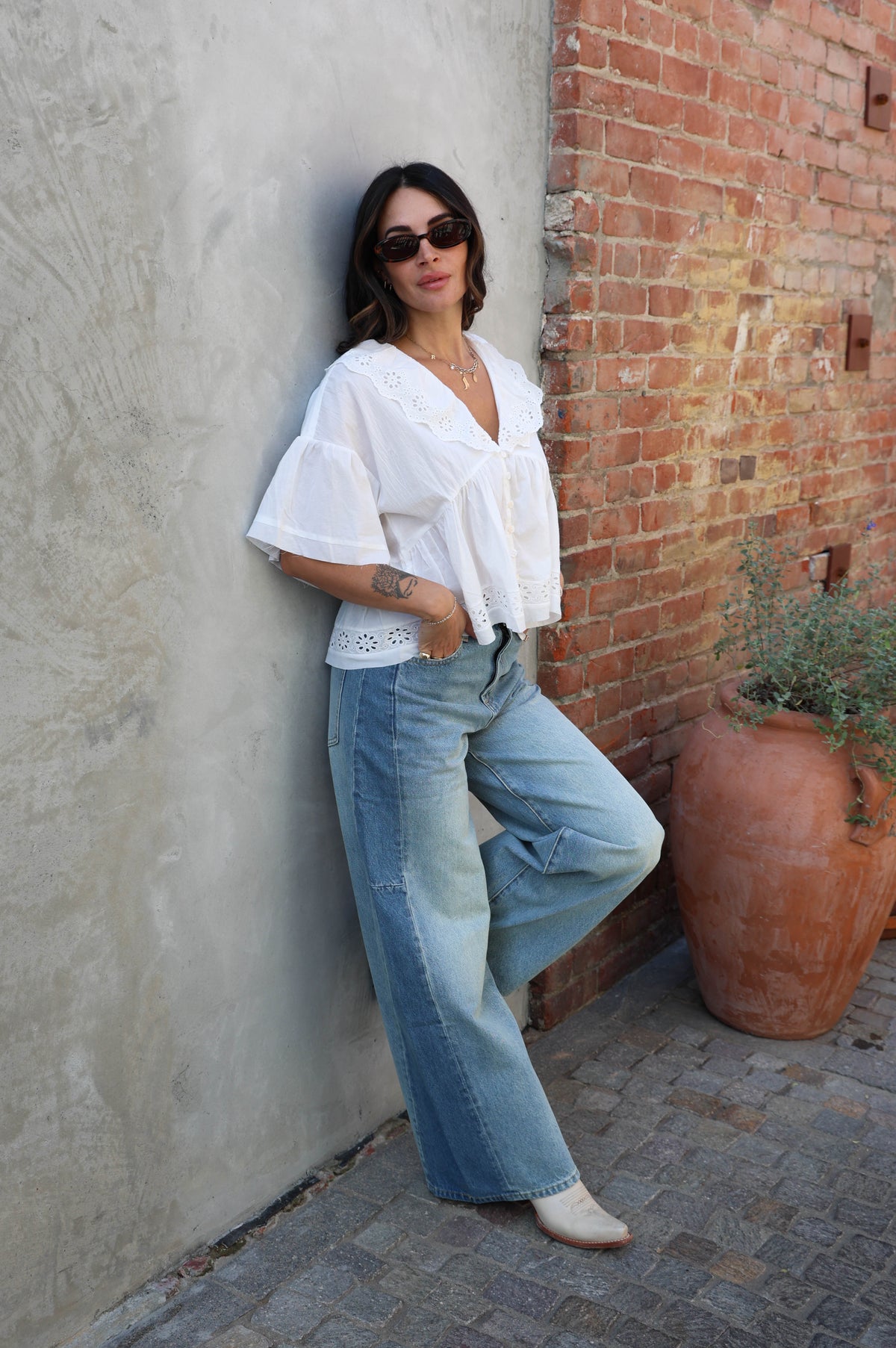 Woman leaning against a brick wall wearing a white blouse and blue jeans.