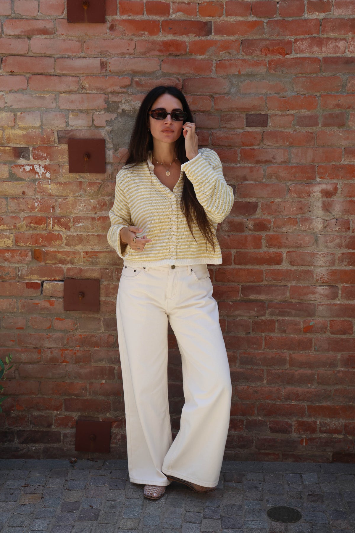 Woman in a yellow striped shirt and white pants standing against a brick wall.