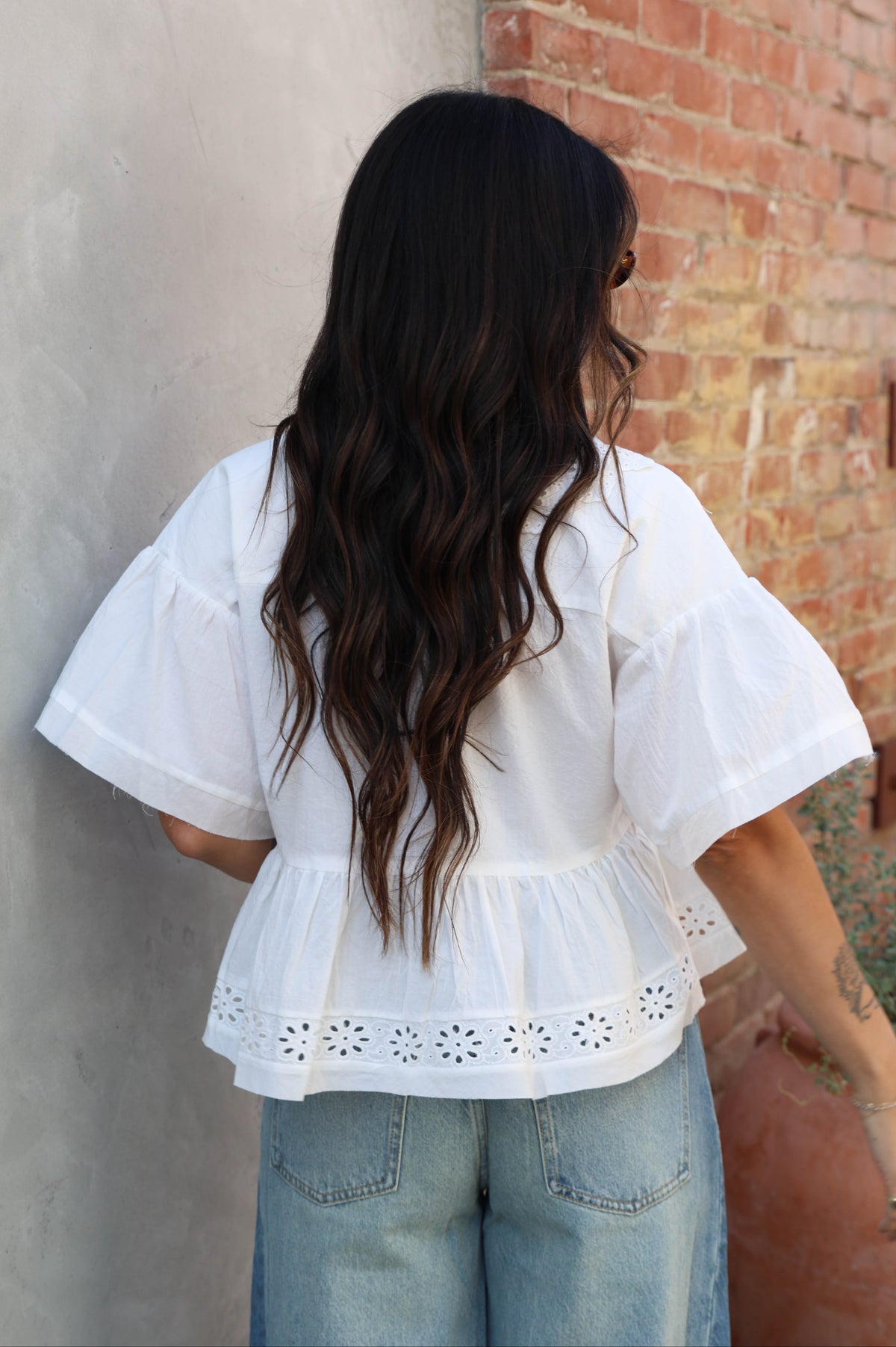 Woman wearing a white blouse with ruffled sleeves and blue jeans against a brick wall.
