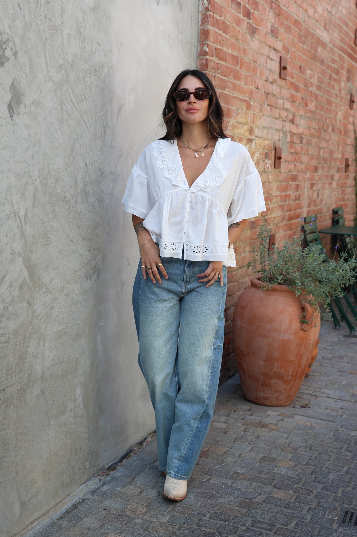 Woman in a white blouse and blue jeans standing against a textured wall with a large terracotta pot nearby.