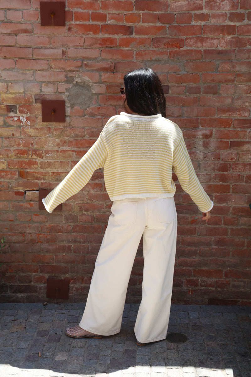 Person wearing a yellow striped sweater and white pants standing against a brick wall.