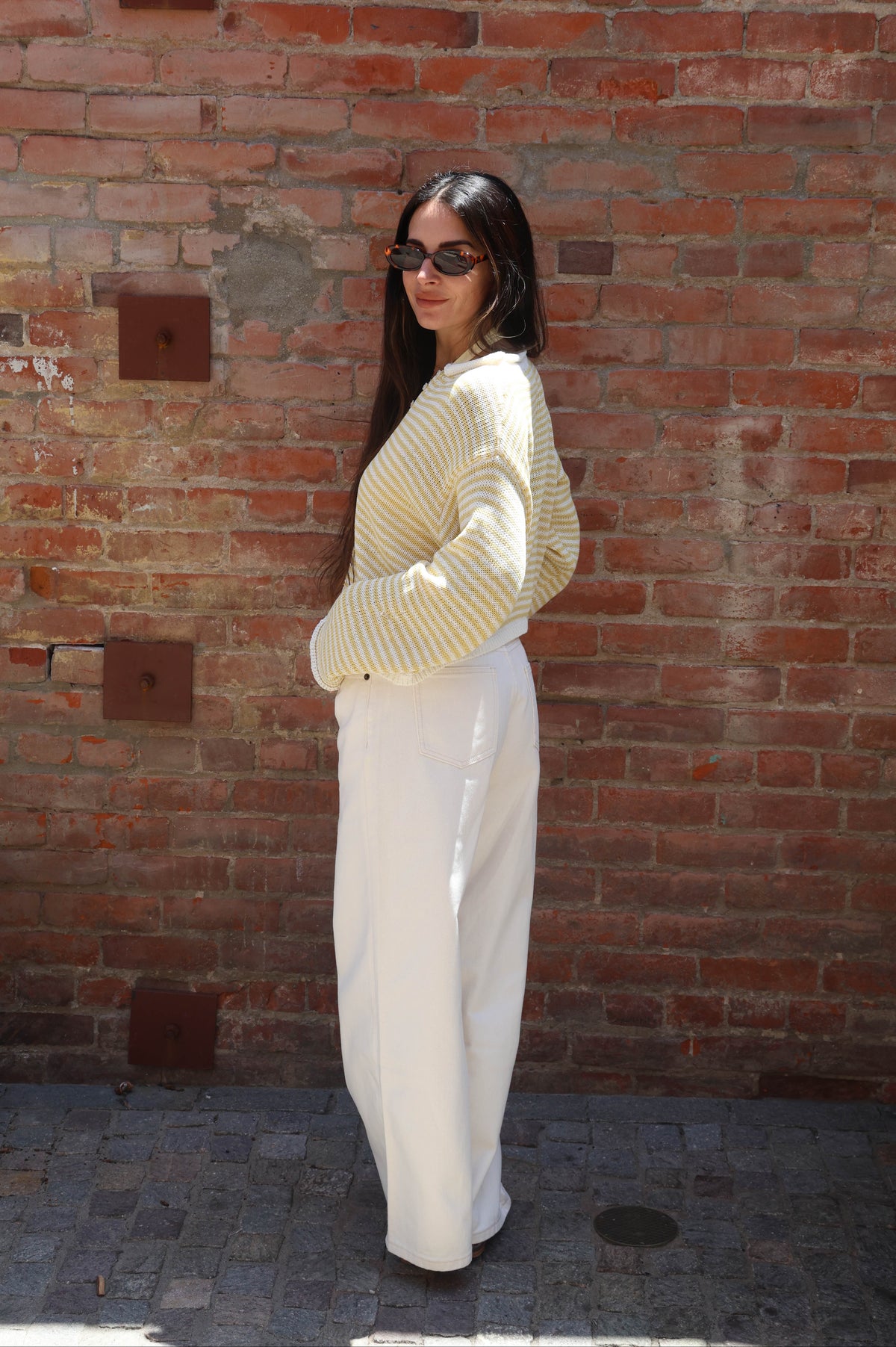 Woman wearing a striped sweater and white pants standing against a brick wall.
