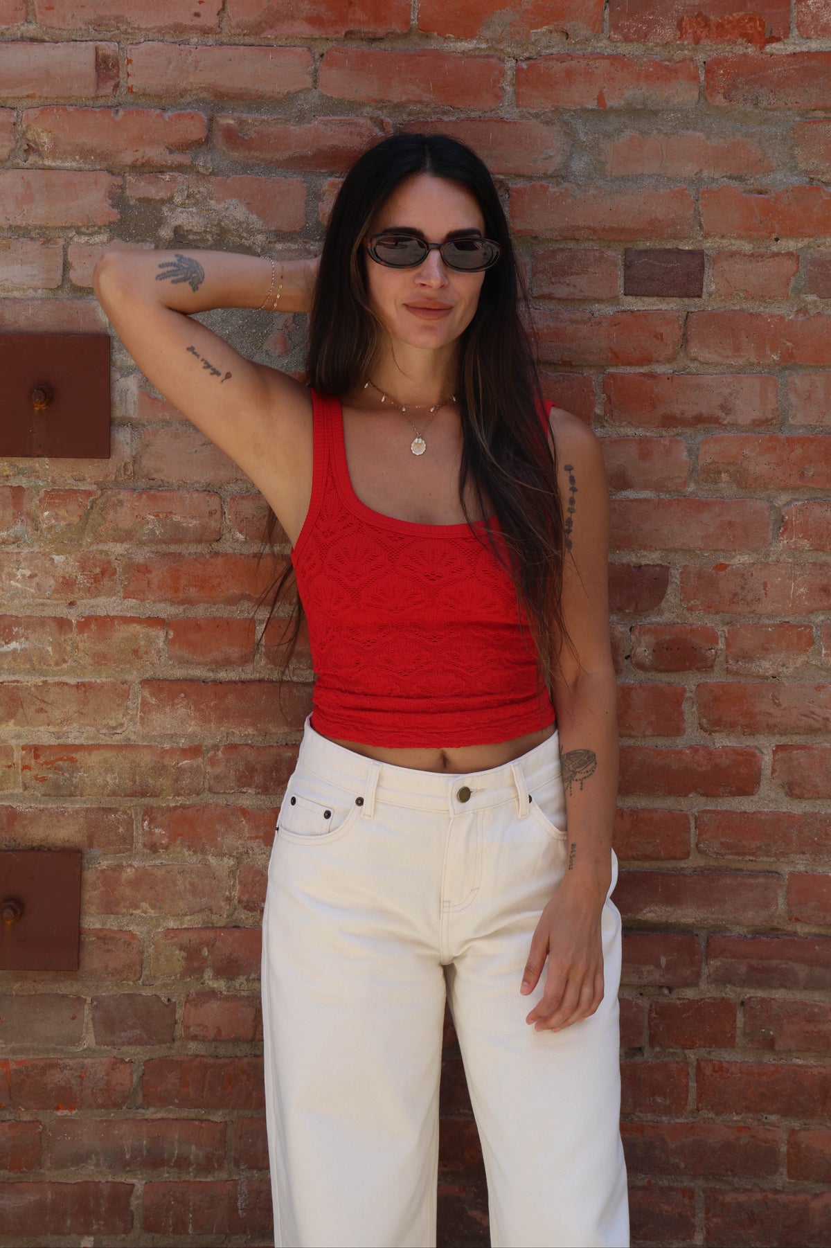 Woman wearing a red top and white pants standing against a brick wall.