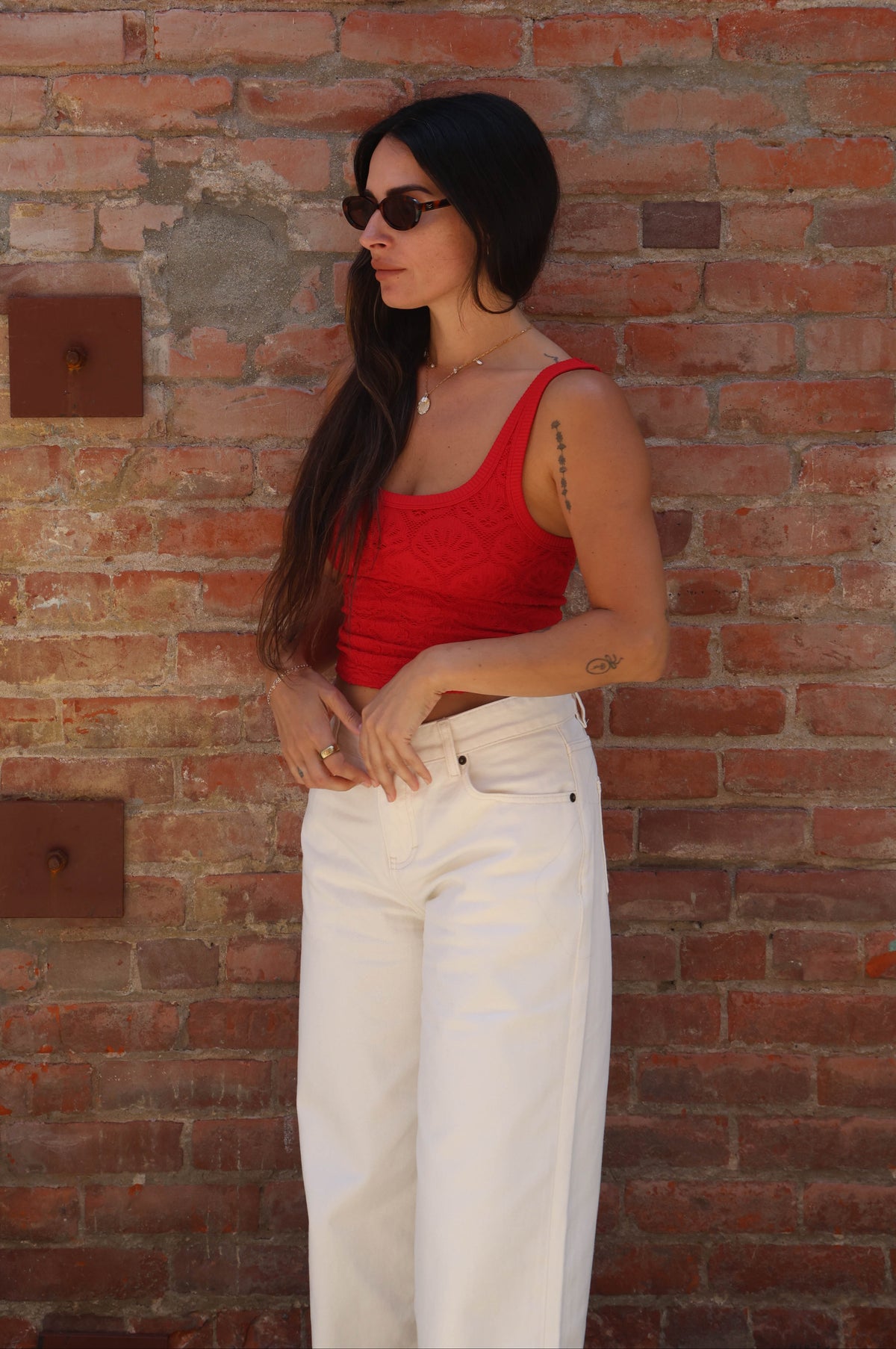 Woman wearing a red tank top and white pants against a brick wall.