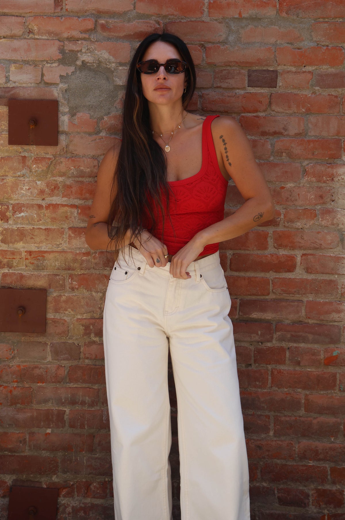 Woman in a red top and white pants standing against a brick wall.