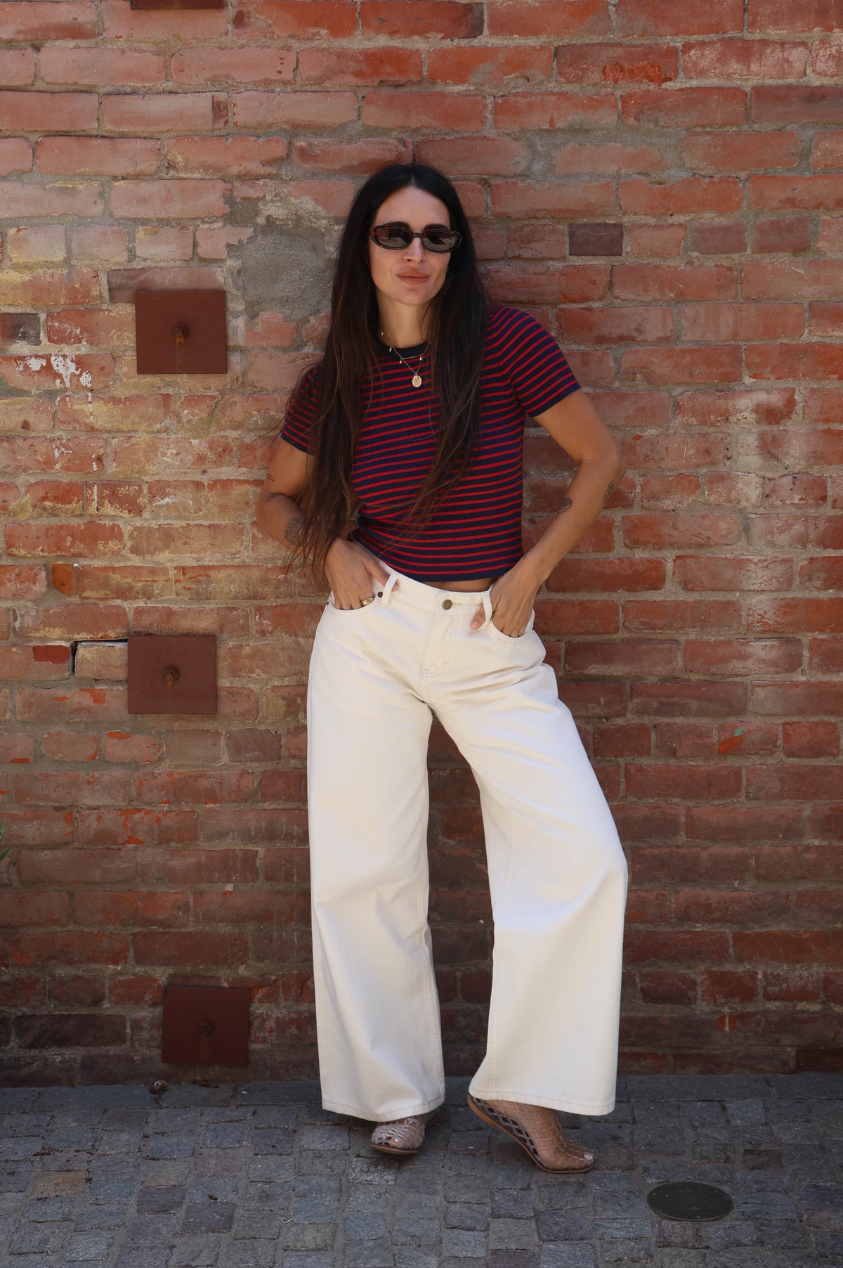 Person wearing a striped shirt and white pants standing against a brick wall.