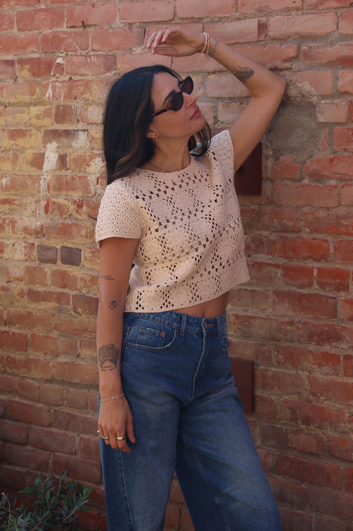 Woman wearing a white lace top and blue jeans against a brick wall.