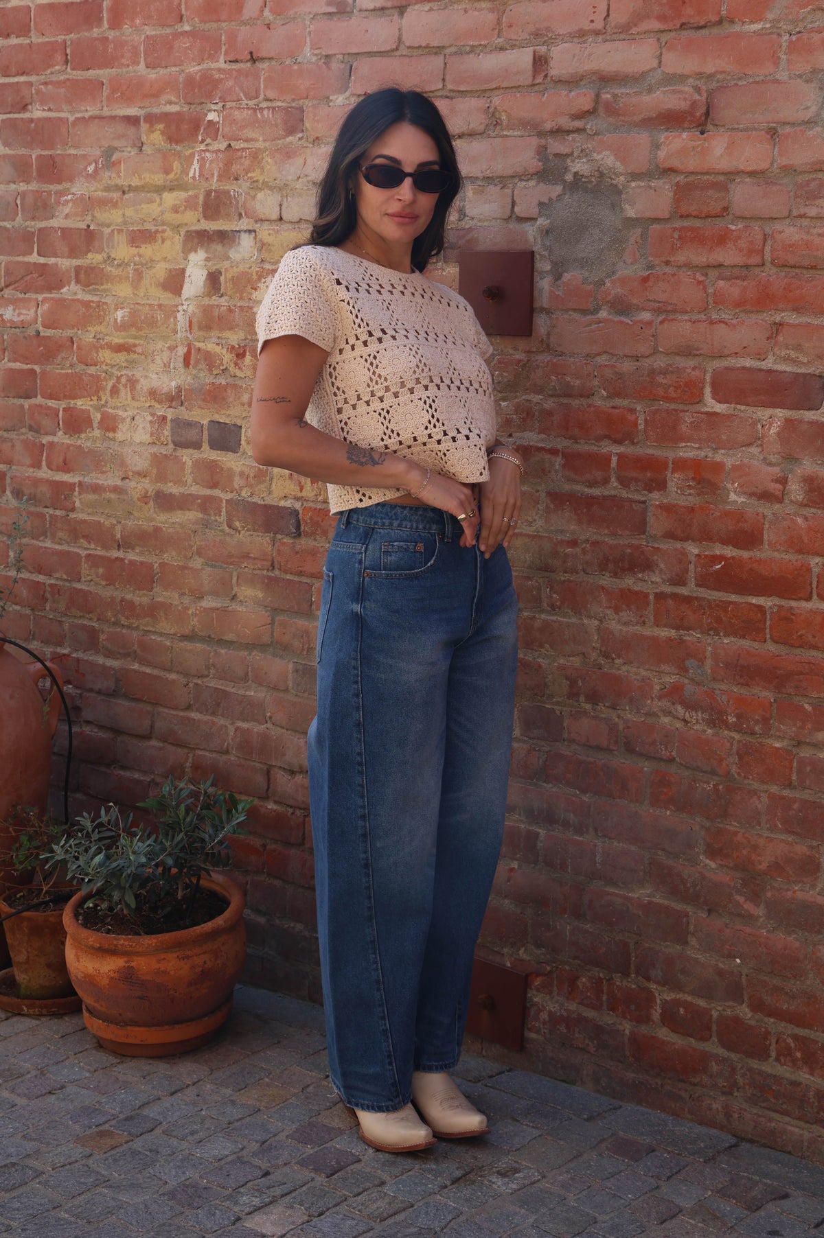 Woman standing against a brick wall wearing sunglasses and casual clothing.