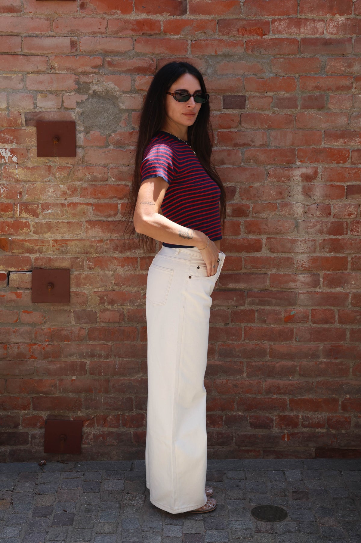Woman wearing a striped shirt and white pants standing against a brick wall.