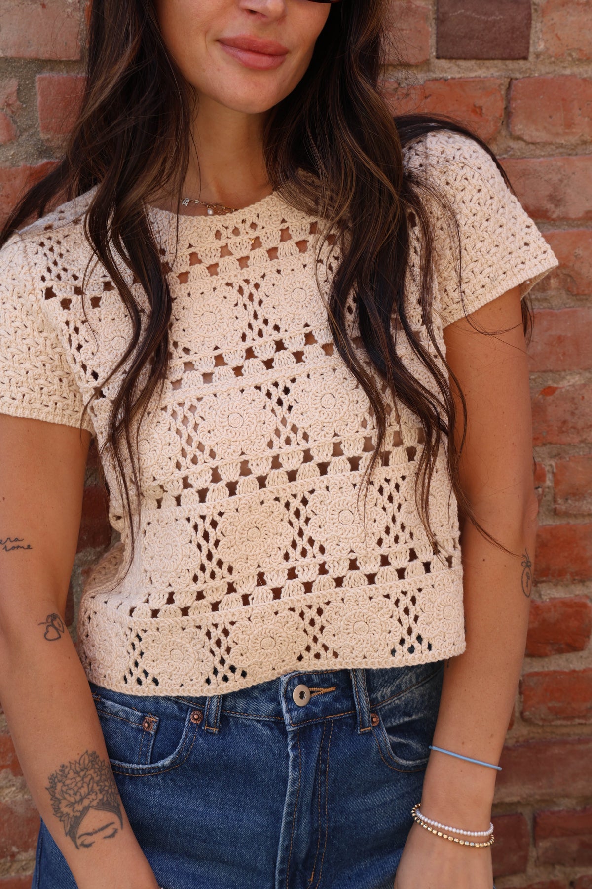 Woman wearing a beige crochet top and blue jeans against a brick wall.